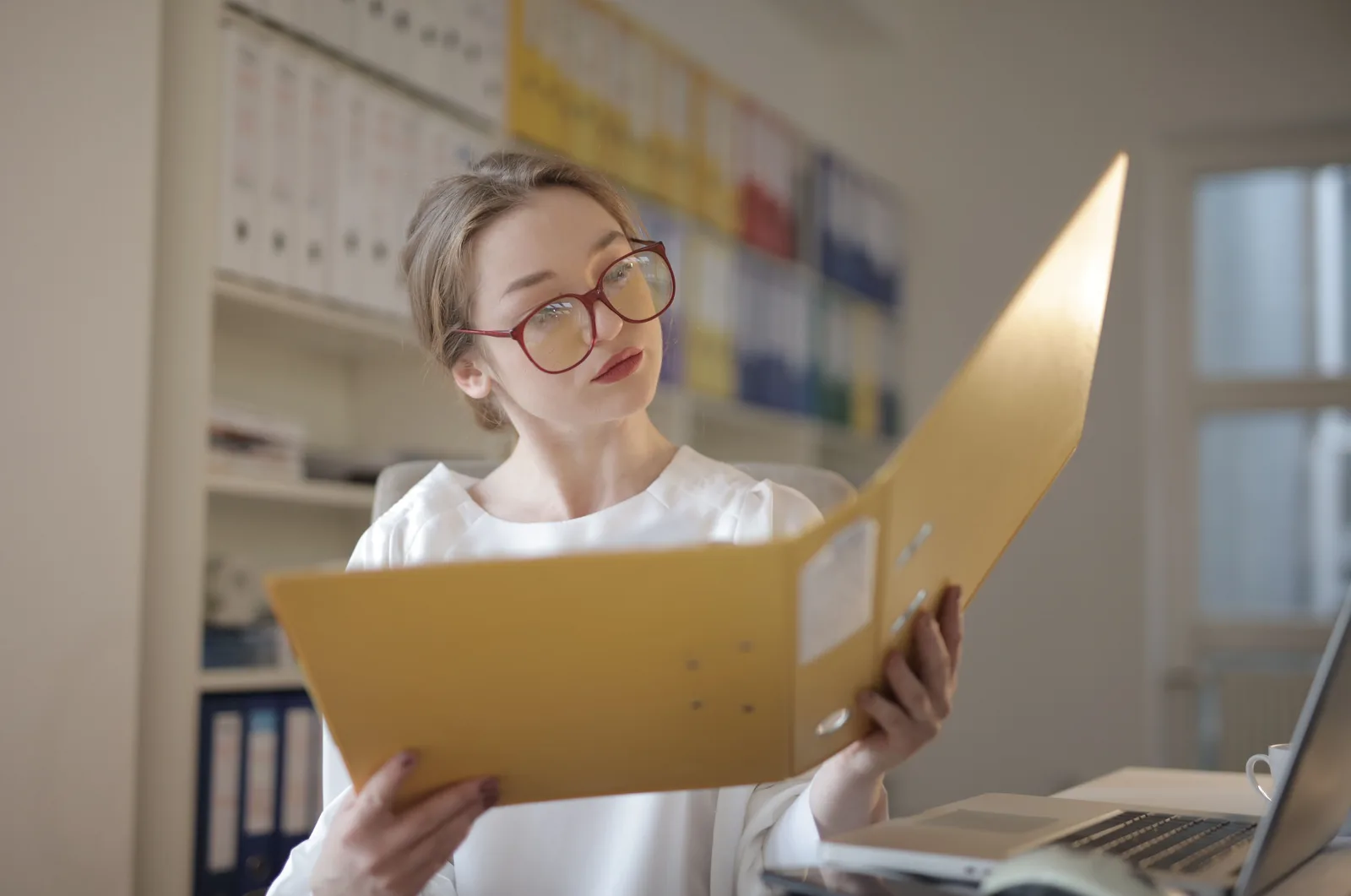An accountant in glasses reading an open client folder at a desk, with a wall of colour-coded binders behind