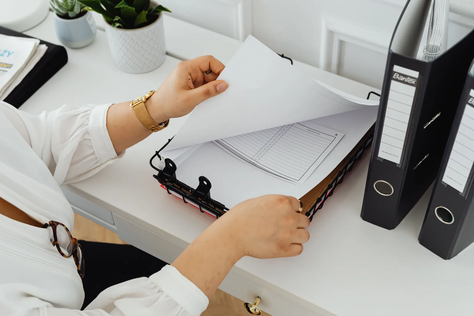 Hands turning pages in a ring binder on a white desk beside two black lever-arch files