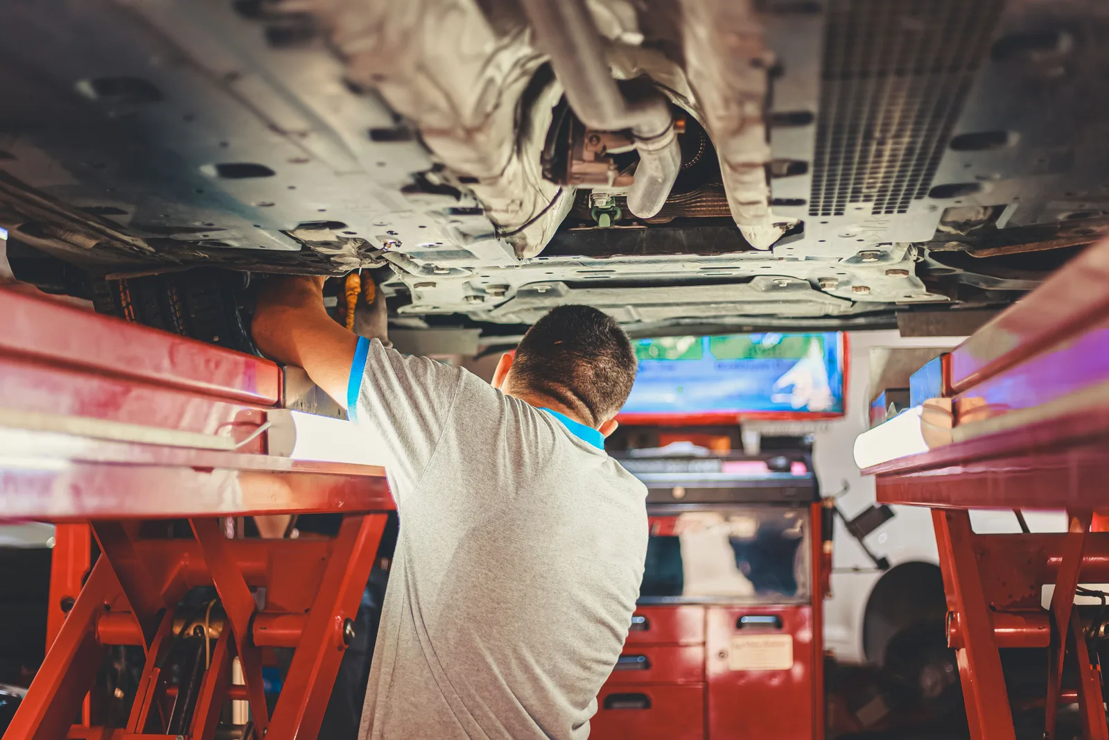 A technician standing under a car raised on a red two post lift, reaching up into the underbody with a rolling toolbox behind him