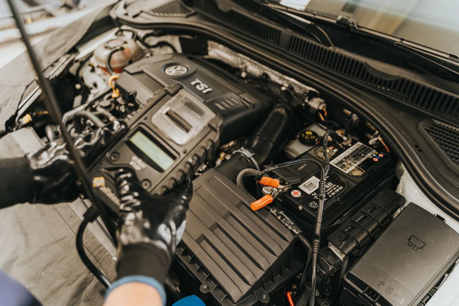 Gloved hands working in an open engine bay with the hood raised, battery leads clipped on and the air box and fuse box exposed
