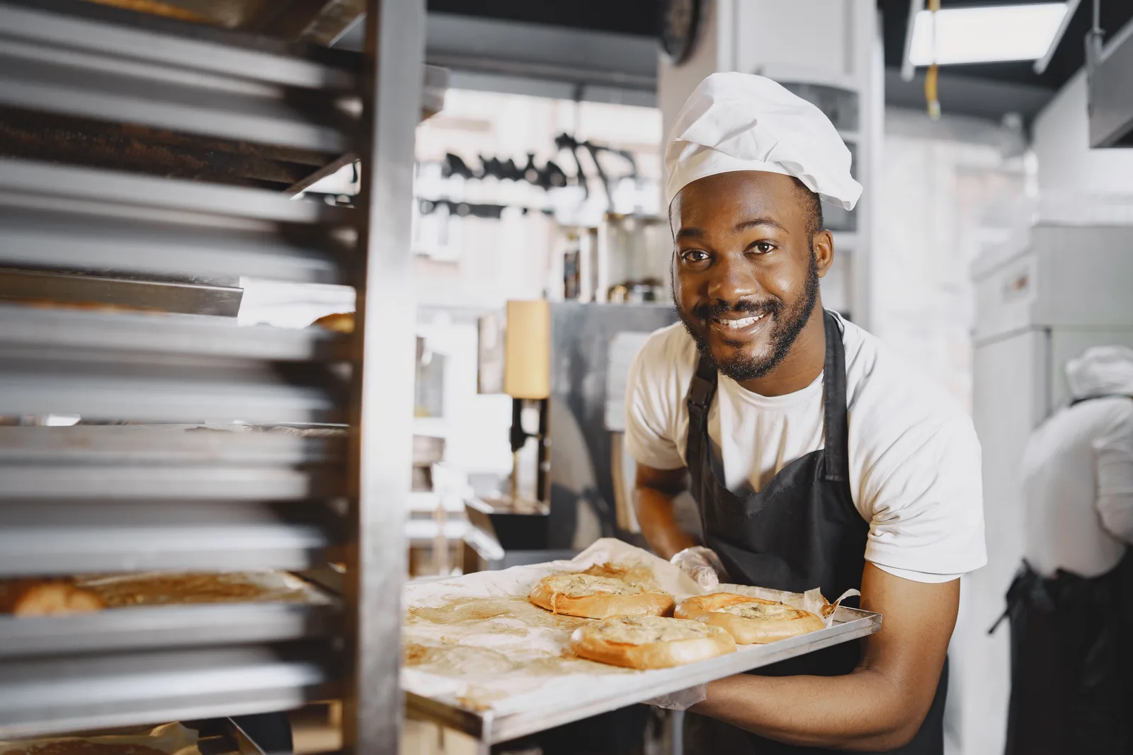 A baker in an apron and chef's hat carrying a tray of bread rolls beside a tall bakery rack oven
