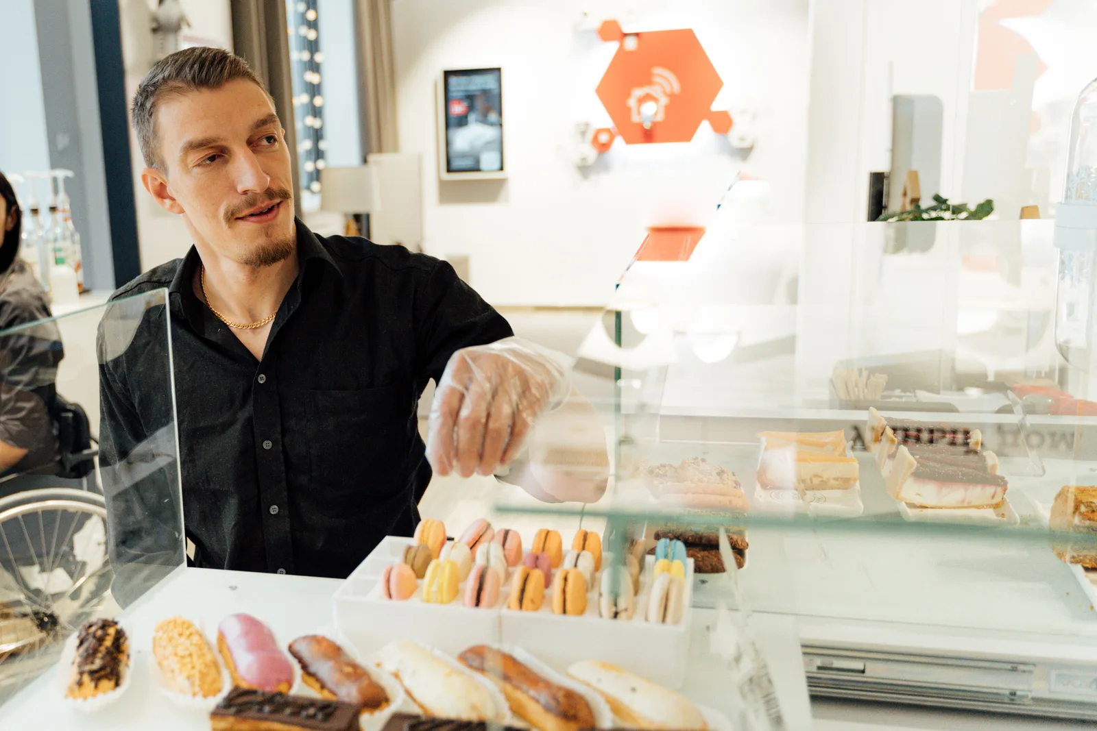A pastry chef wearing gloves arranging eclairs and macarons in a bakery display case