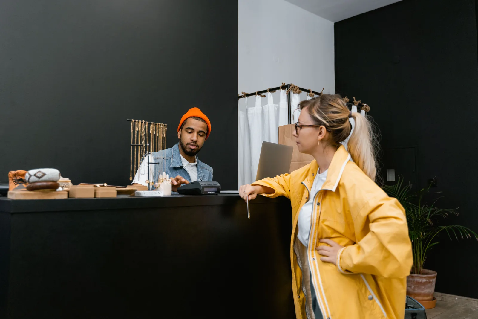 A customer at the counter of a small clothing and jewellery shop while an assistant works the card terminal behind it