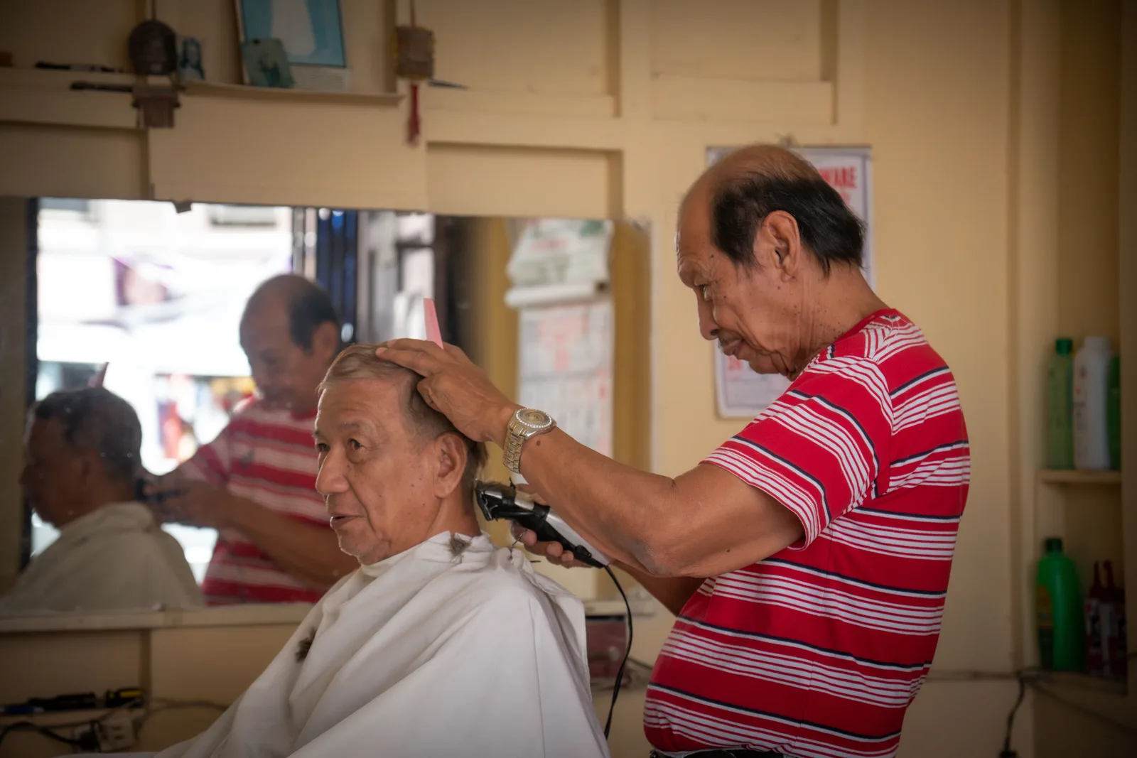 Barber using clippers on an older man’s hair while a second barber works behind him