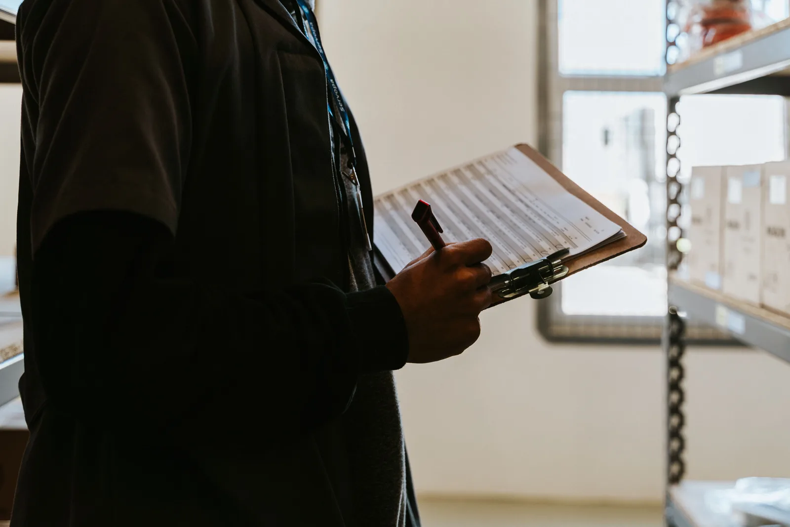 A worker holding a pen over a printed form on a clipboard in a warehouse, with racked stock behind.