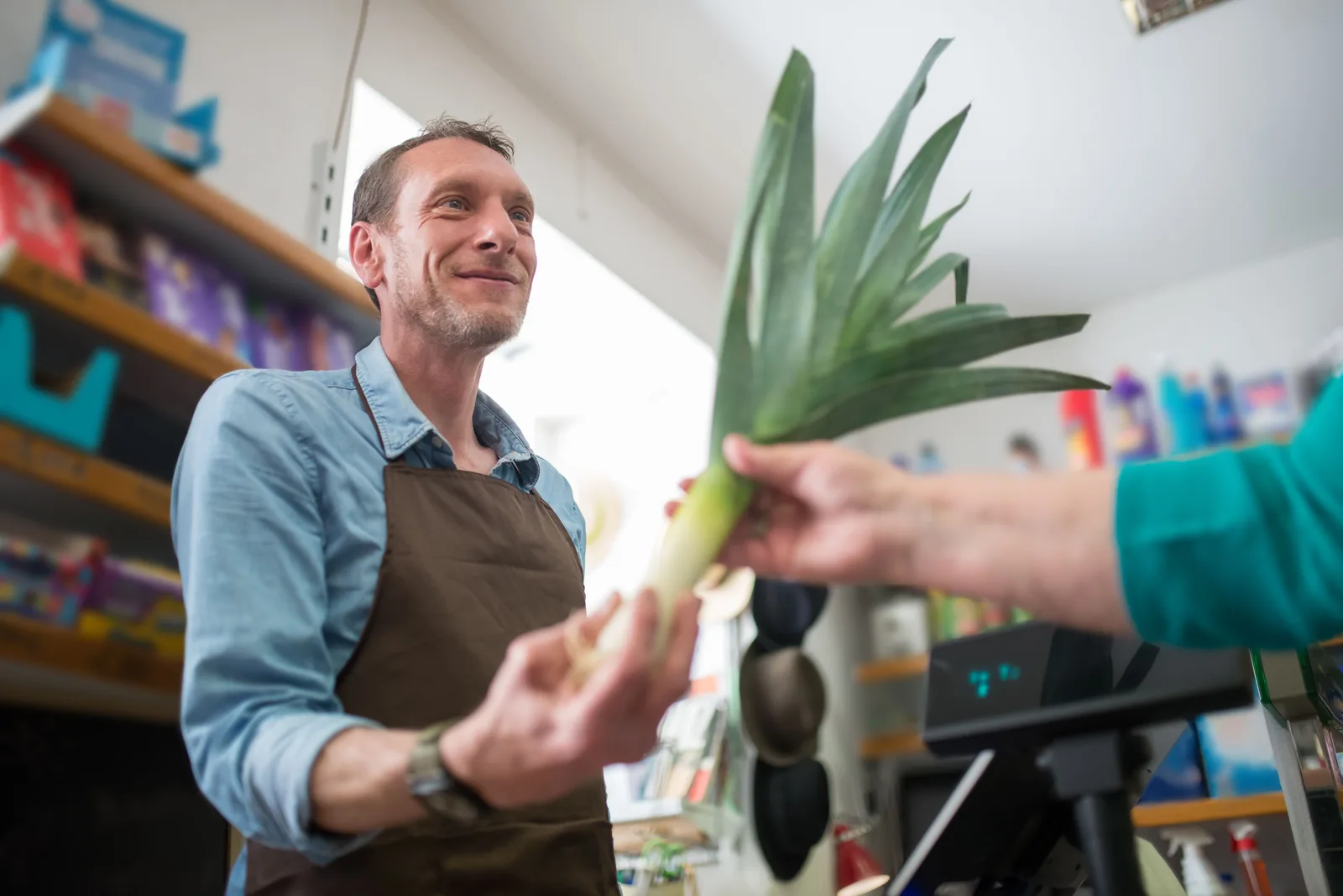 A shopkeeper in an apron handing a leek across the counter to a customer, with a card terminal beside the till