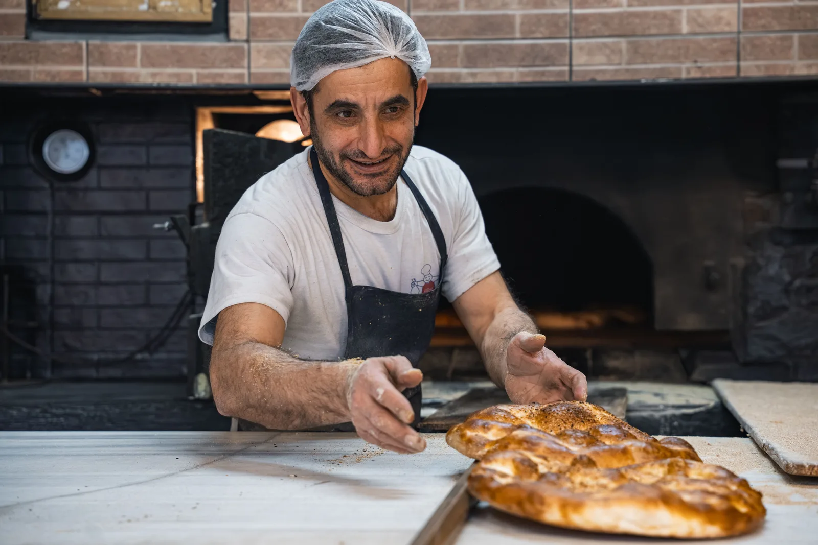 A baker in an apron and hairnet sliding a large flatbread off a peel onto the counter beside a brick oven.