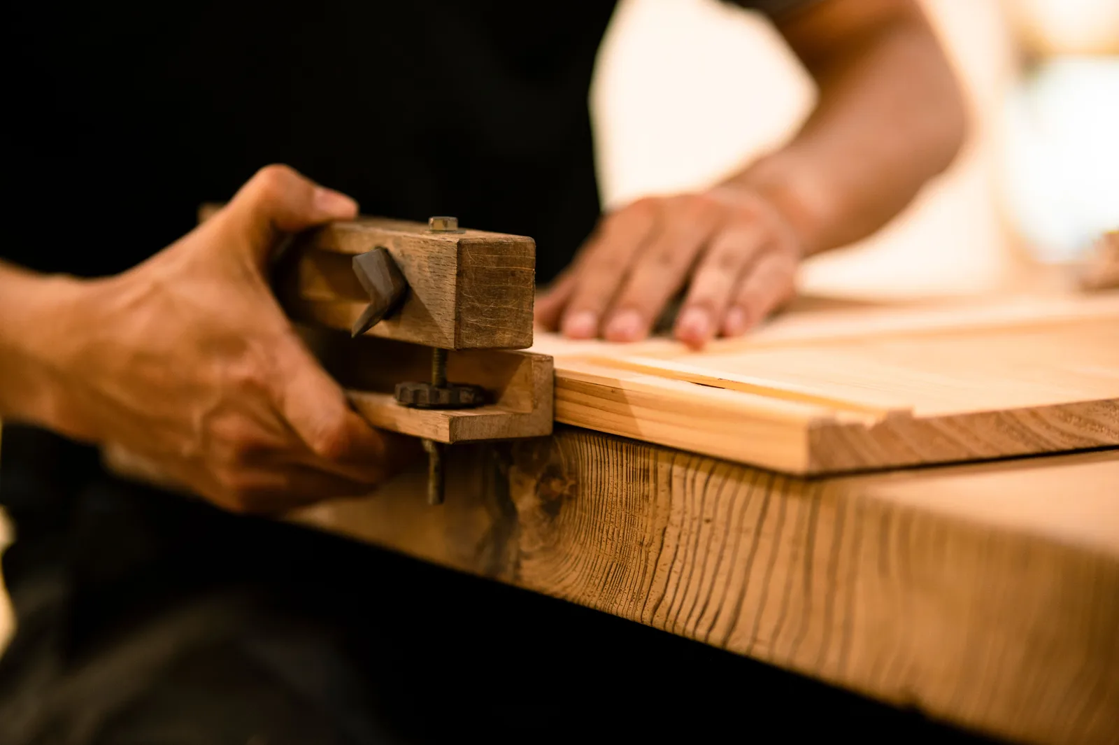 Close view of a woodworker hands running a hand plane along the edge of a board on a workbench.