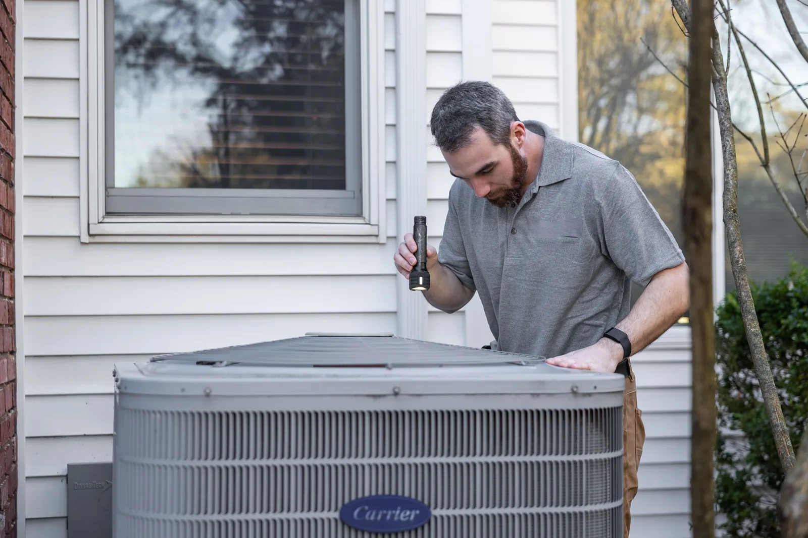 A technician holding a torch light and leaning over an outdoor air conditioning condenser beside a house