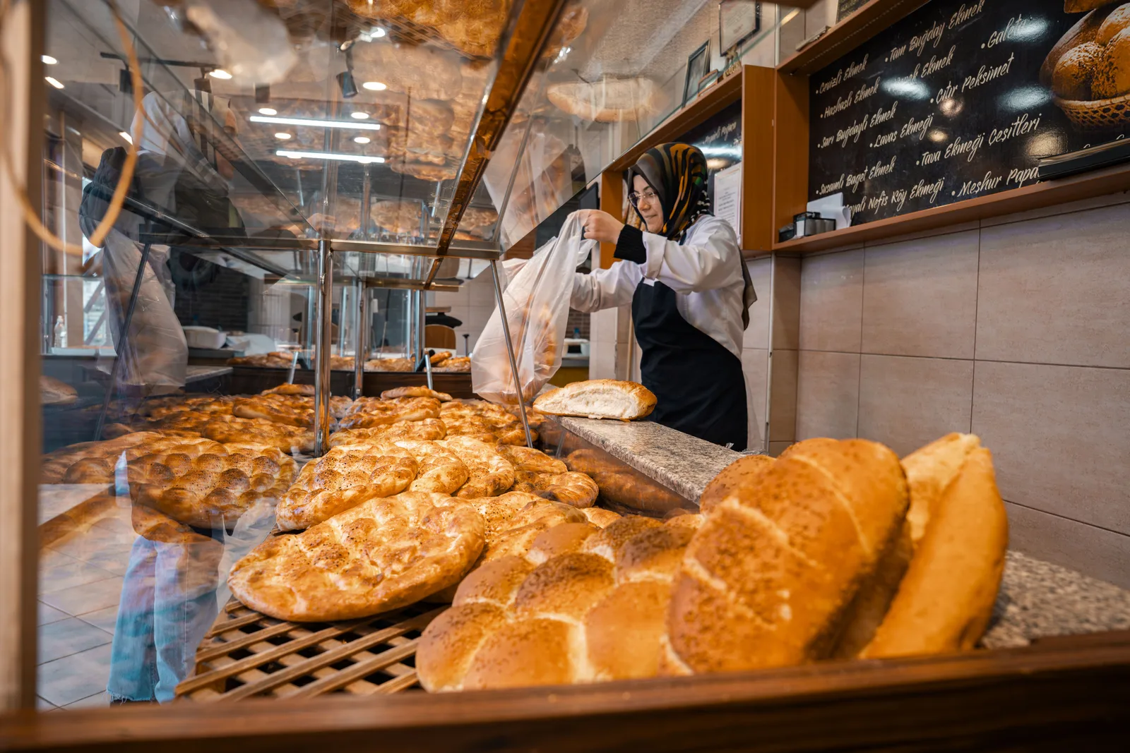 A baker in a white coat bagging bread behind a bakery counter stacked with fresh loaves and pastries