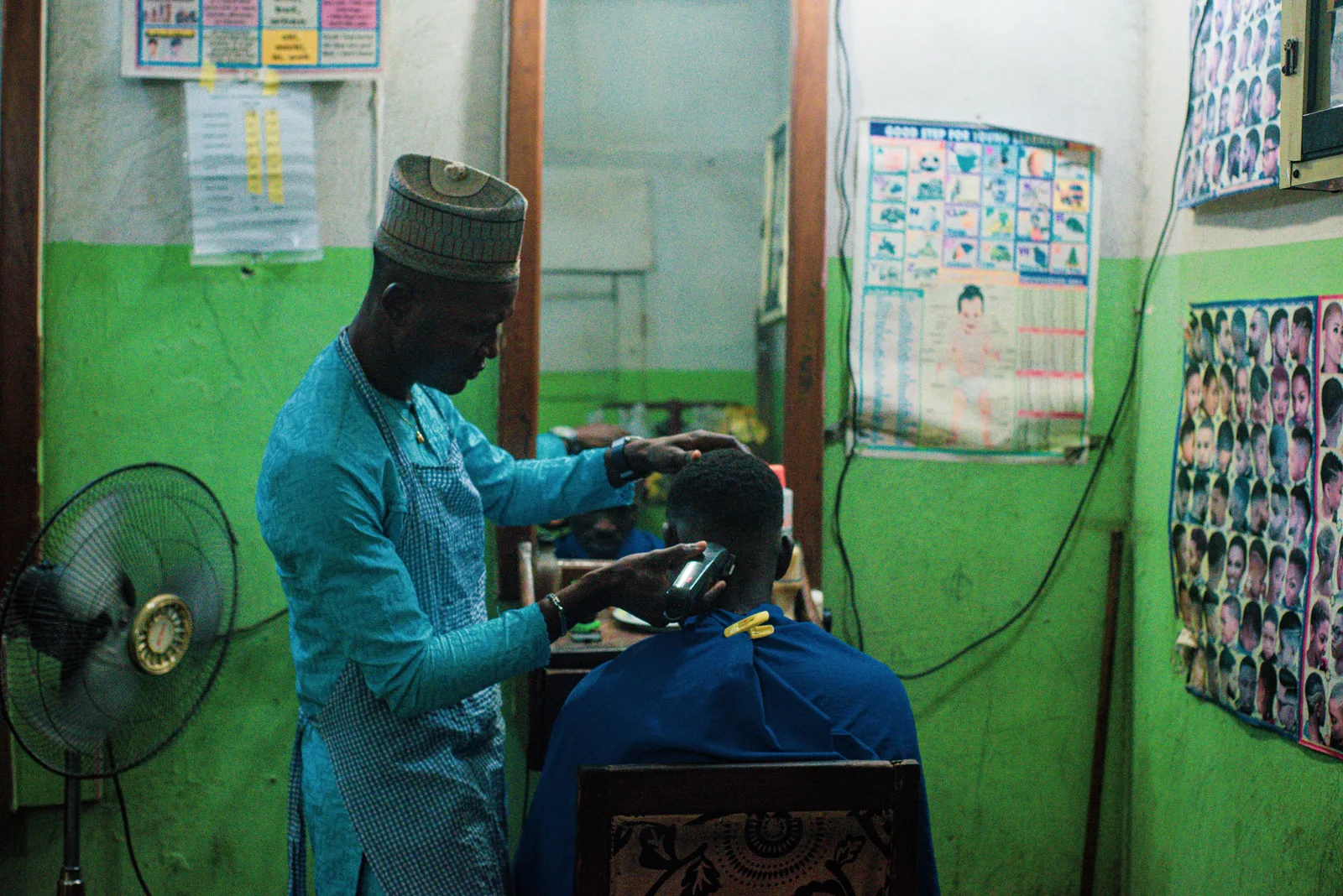 A barber running clippers through a seated customer's hair in a traditional barbershop, with a second chair and mirrors behind