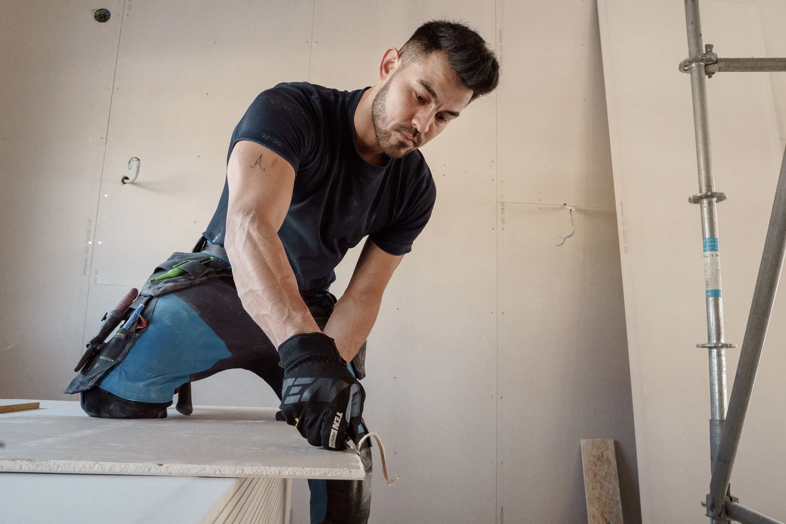 Un trabajador con guantes cortando una lámina de tablarroca dentro de un edificio en construcción.