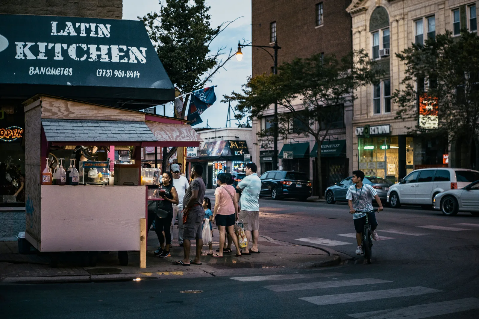 Customers waiting at a Latin Kitchen food cart on a Chicago sidewalk in the evening, with lit storefronts behind