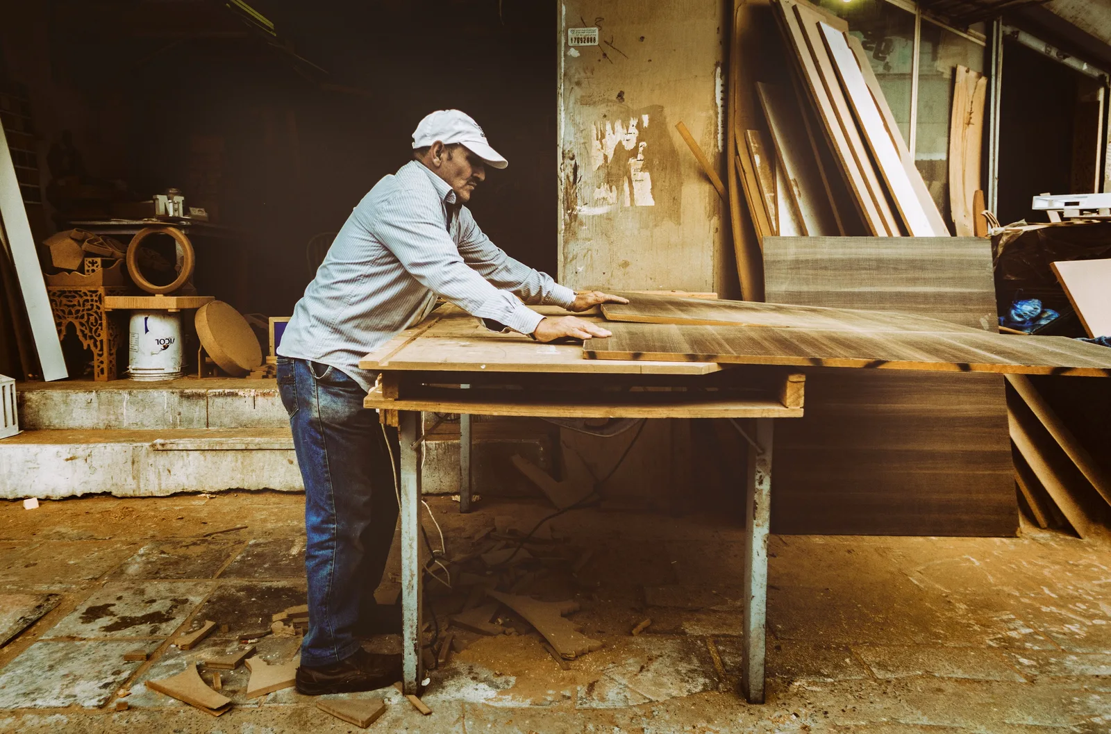 A carpenter working a long board across a bench in a timber workshop, with lumber stacked against the wall behind him.