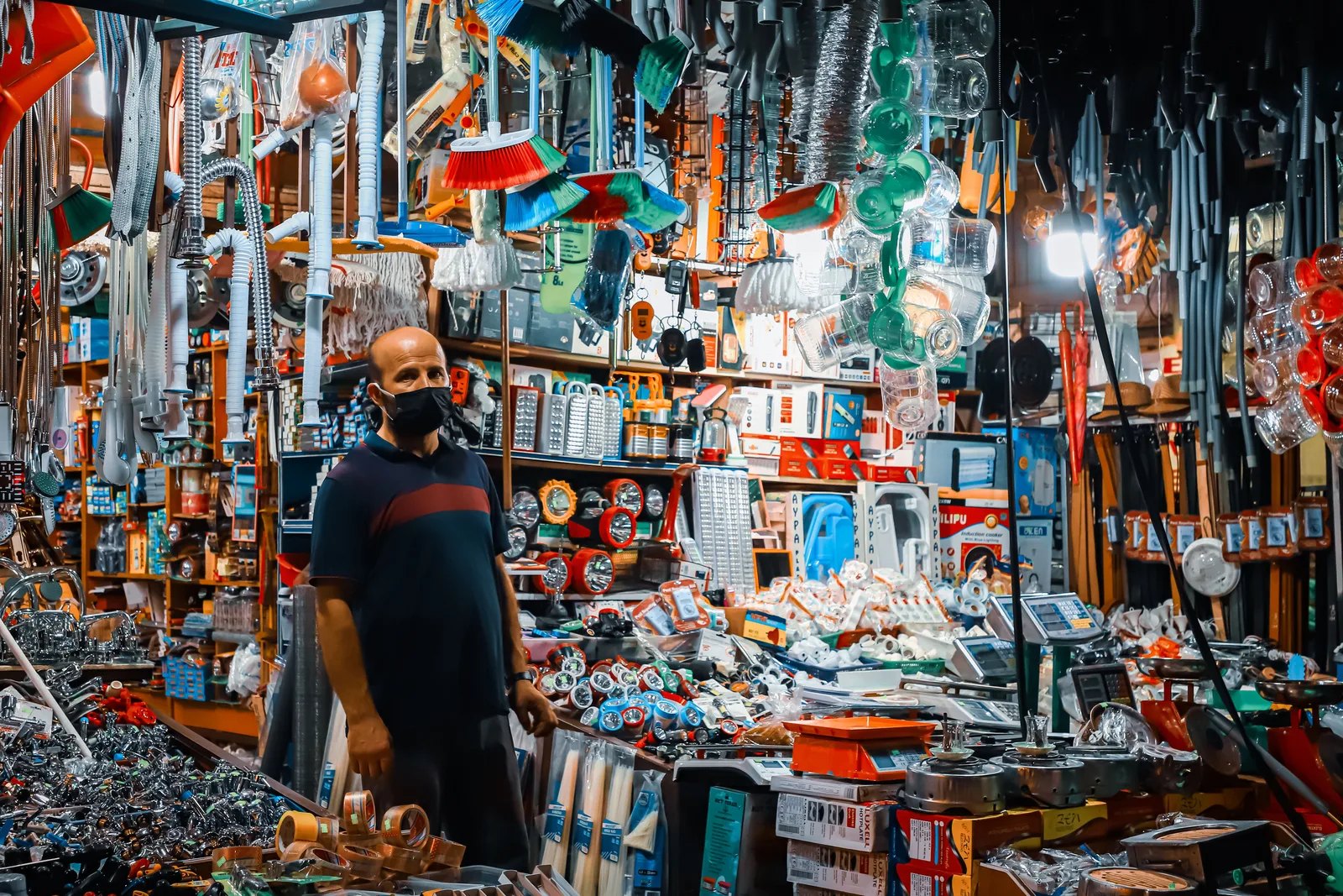 A shopkeeper standing in a narrow aisle of a hardware and houseware store packed floor to ceiling with stock.