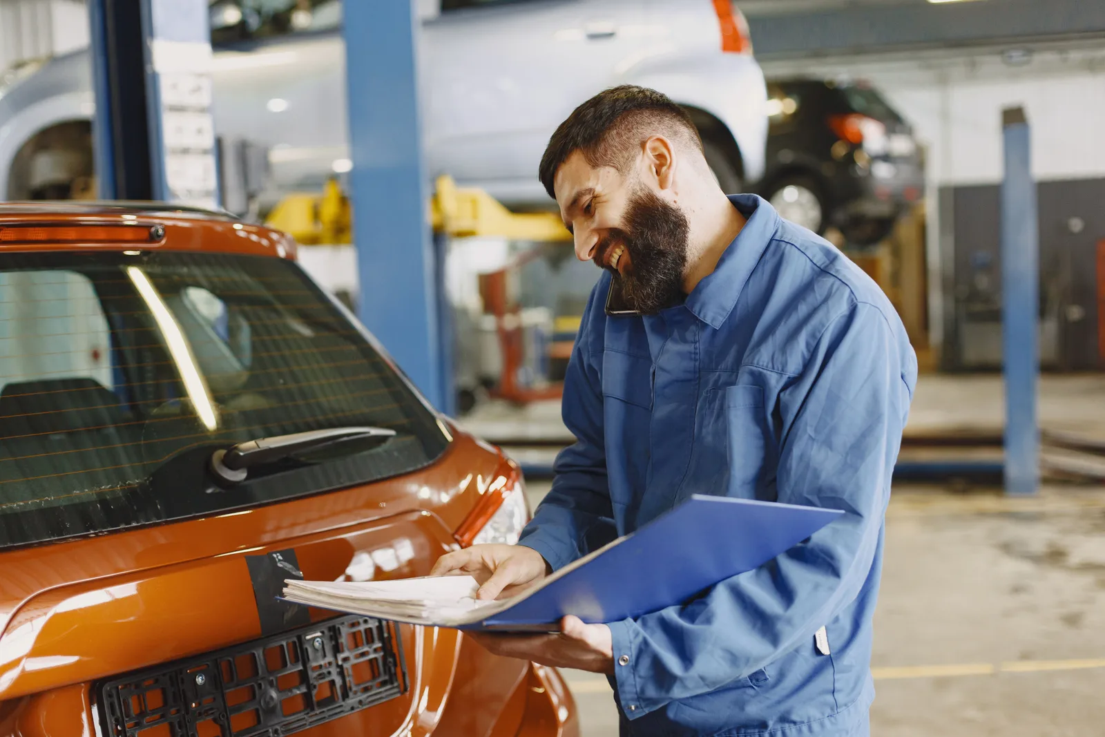 Mechanic in coveralls writing on a clipboard beside a car in his own workshop.