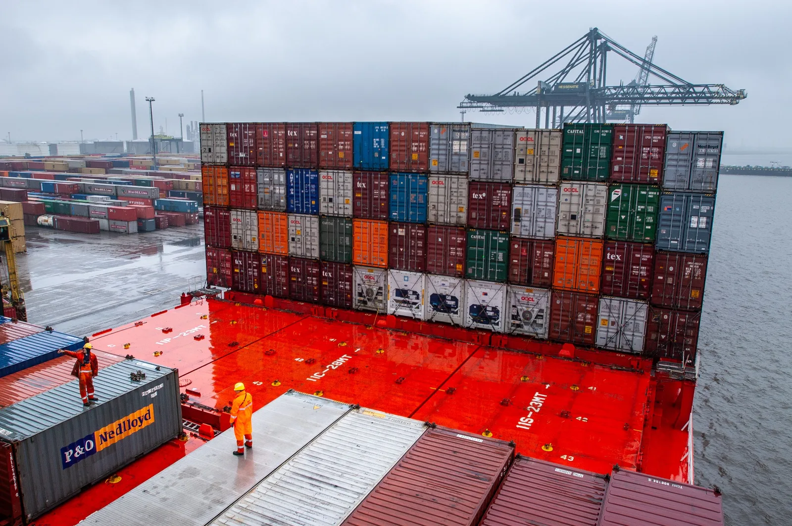 Stacked shipping containers, a moored container vessel and gantry cranes at a container terminal, with a dock worker in orange on the deck below.