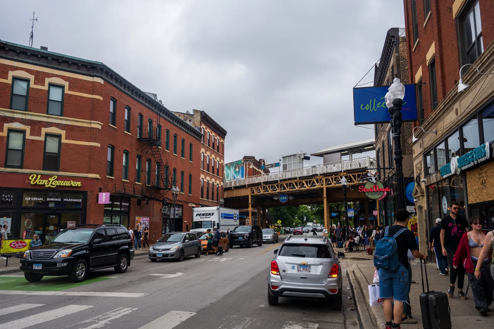 Shops, an ice cream parlor and a donut counter lining a Chicago street beneath the elevated CTA tracks at Damen, with pedestrians on the sidewalk
