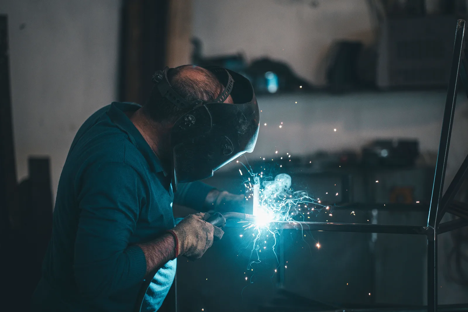 A welder in a protective mask cutting steel on a bench, with sparks scattering across a fabrication workshop.