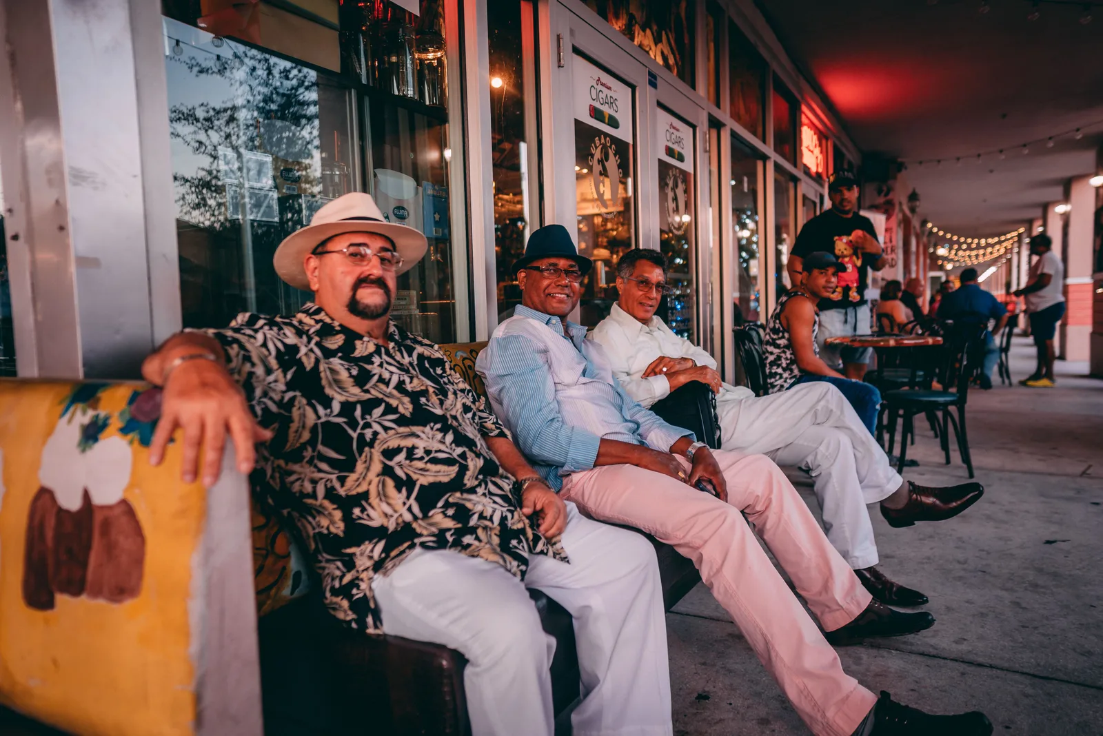 Tres hombres sentados en una banca en la acera afuera de una tabaquería y cafetería de Miami por la noche, con más personas en mesas al aire libre más adelante en la cuadra.