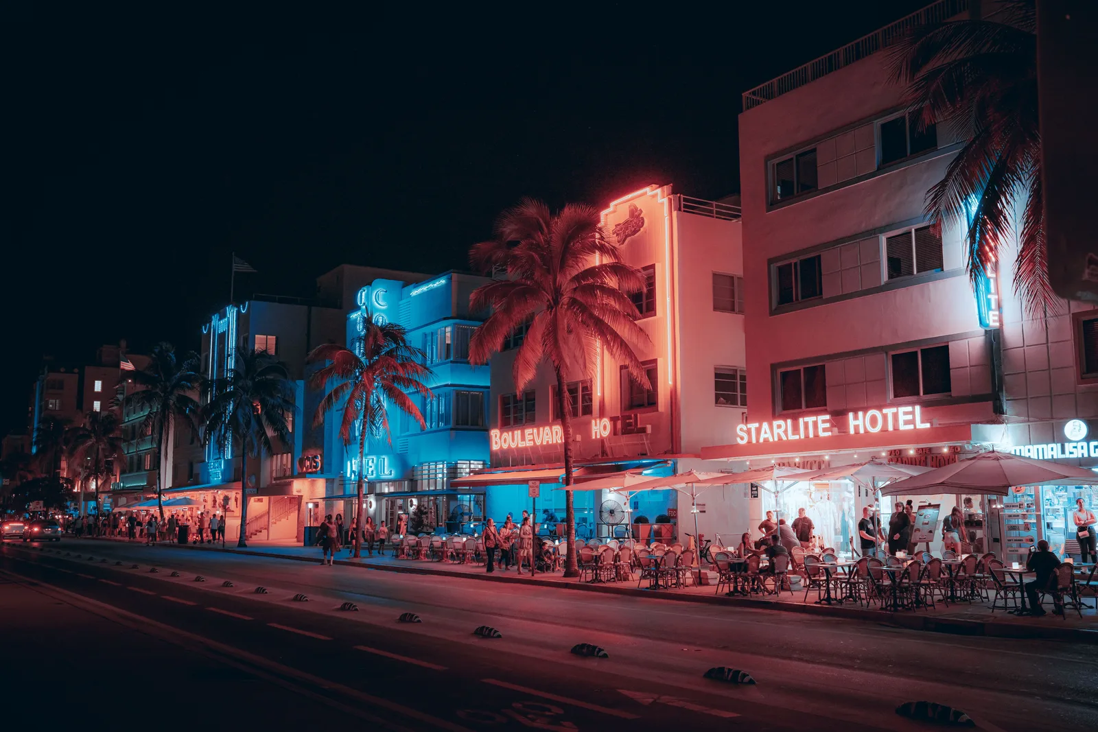 Hoteles y restaurantes estilo Art Deco iluminados a lo largo de Ocean Drive por la noche, con gente caminando y cenando en la calle.