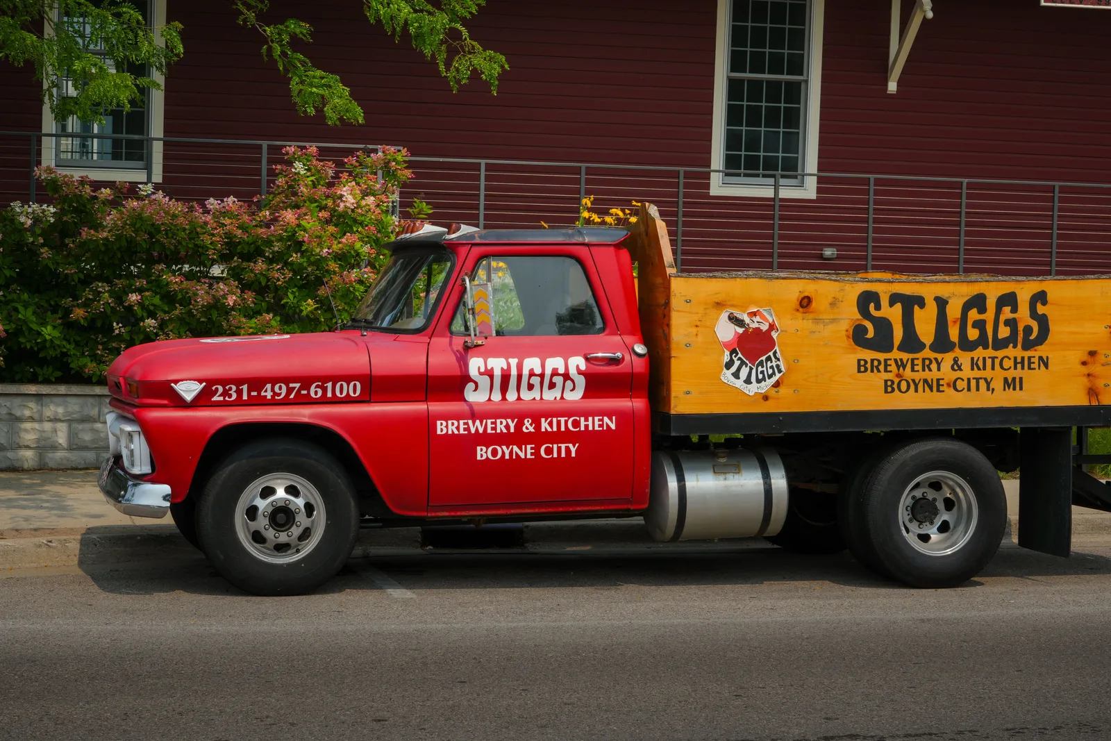 A restored red flatbed truck lettered for a brewery and kitchen parked outside a dark red clapboard building in Boyne City, Michigan