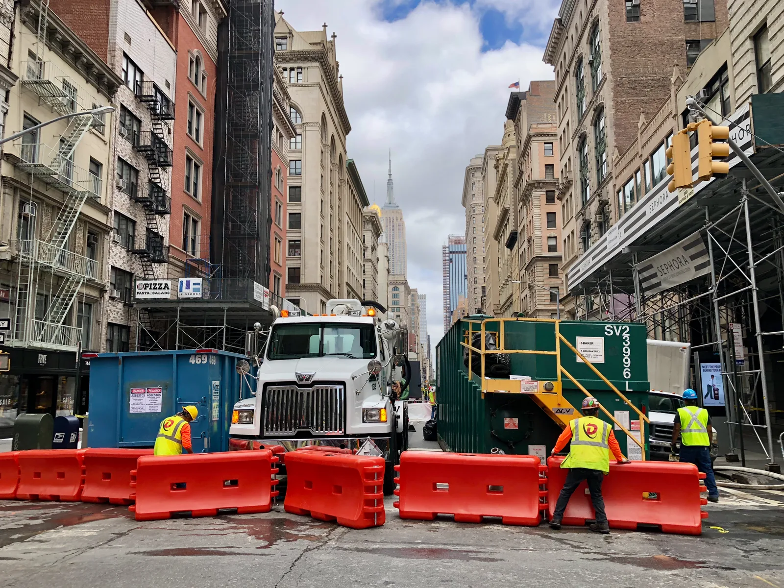Cuadrillas de construcción, un camión volquete y barreras de tráfico anaranjadas ocupando un carril de una avenida de Manhattan bordeada de edificios antiguos.