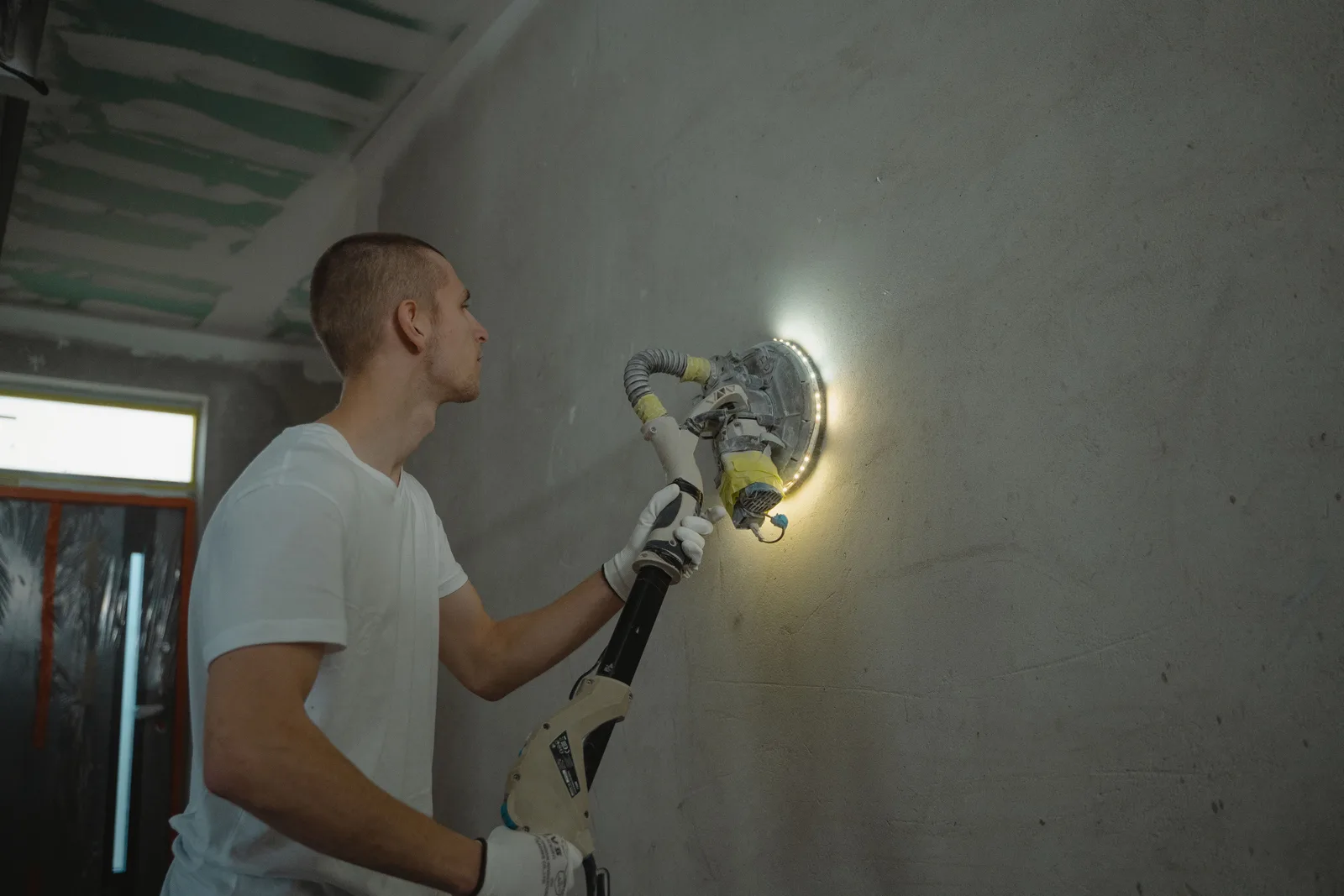 Worker sanding a plaster wall with a power sander during an interior renovation