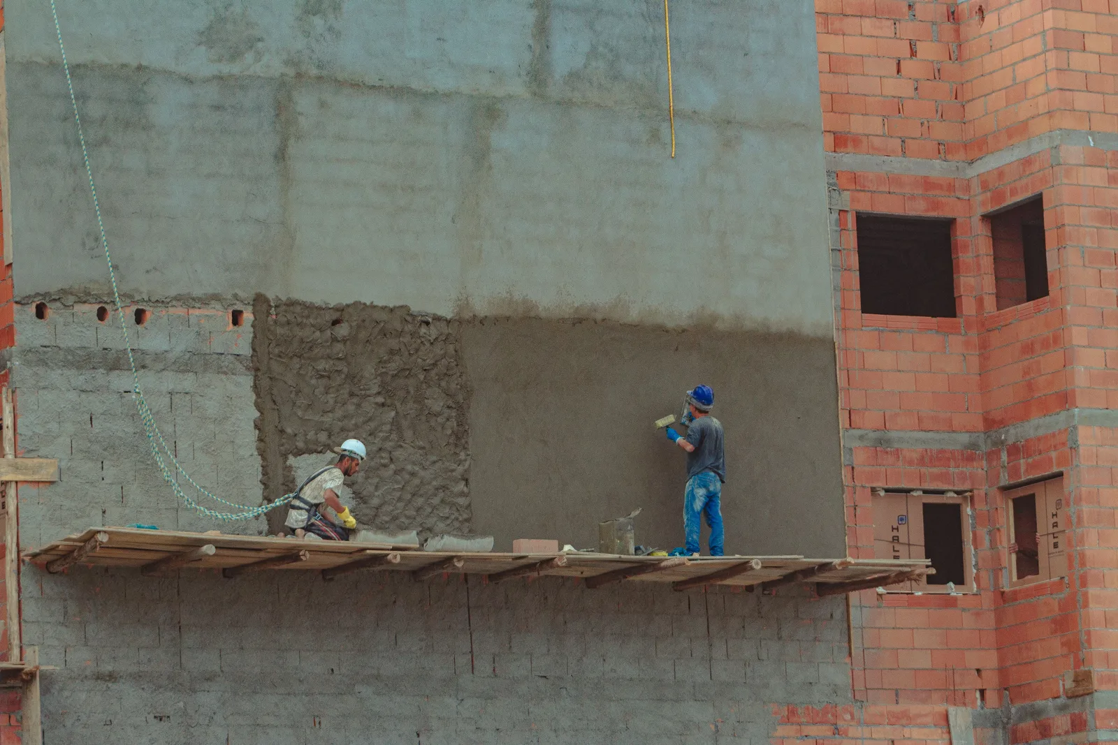 Two workers on a plank scaffold applying plaster to the wall of an unfinished masonry building.