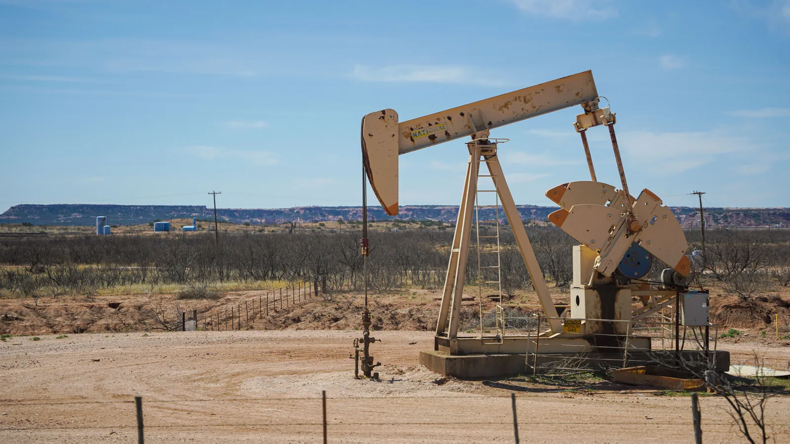 A pumpjack on a fenced well pad in dry West Texas scrub, with a mesa on the horizon.