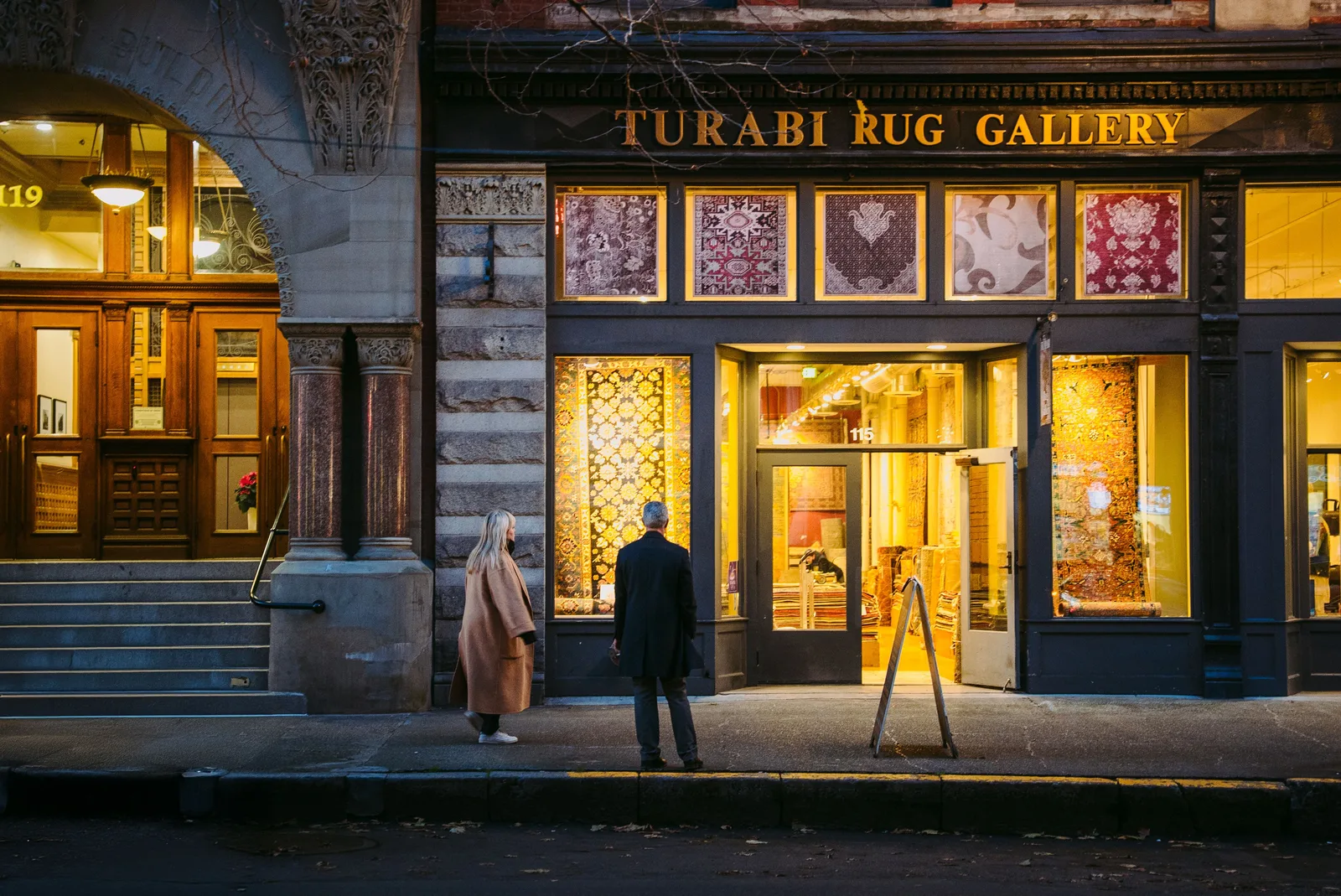 Two people looking into the lit window of a rug gallery on a Seattle street in the evening
