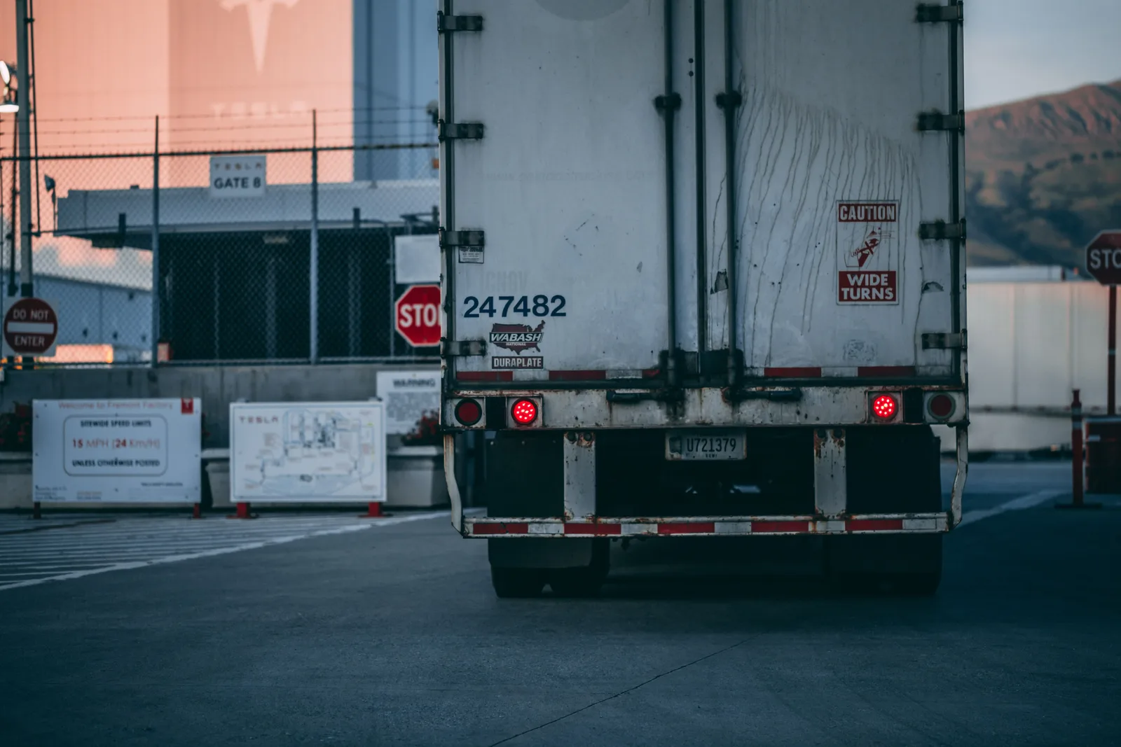 The rear doors of a white freight trailer backed up to a gate at a distribution yard, with a stop sign and fencing alongside.