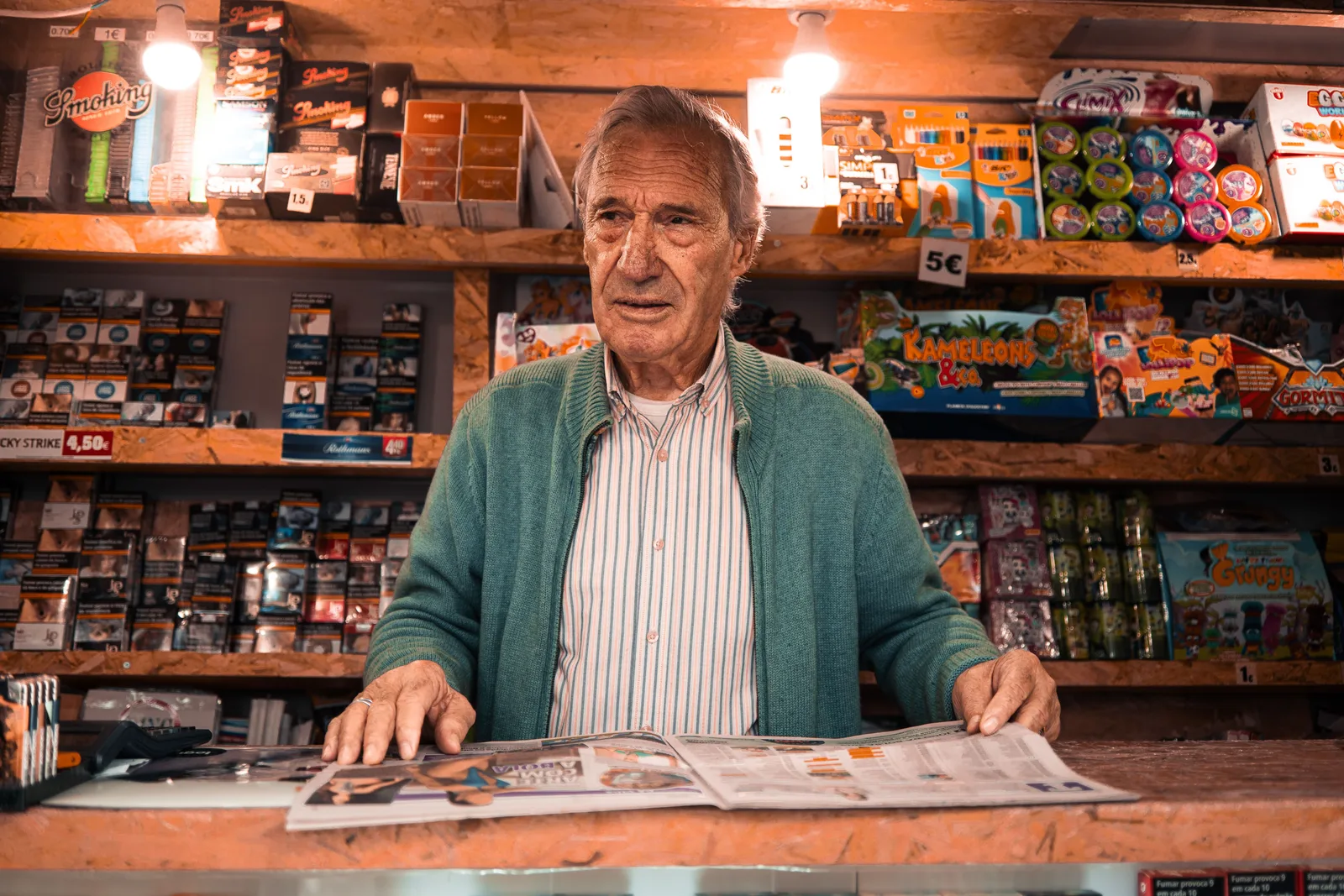 A shopkeeper standing behind the counter of a small newsstand surrounded by stocked products.