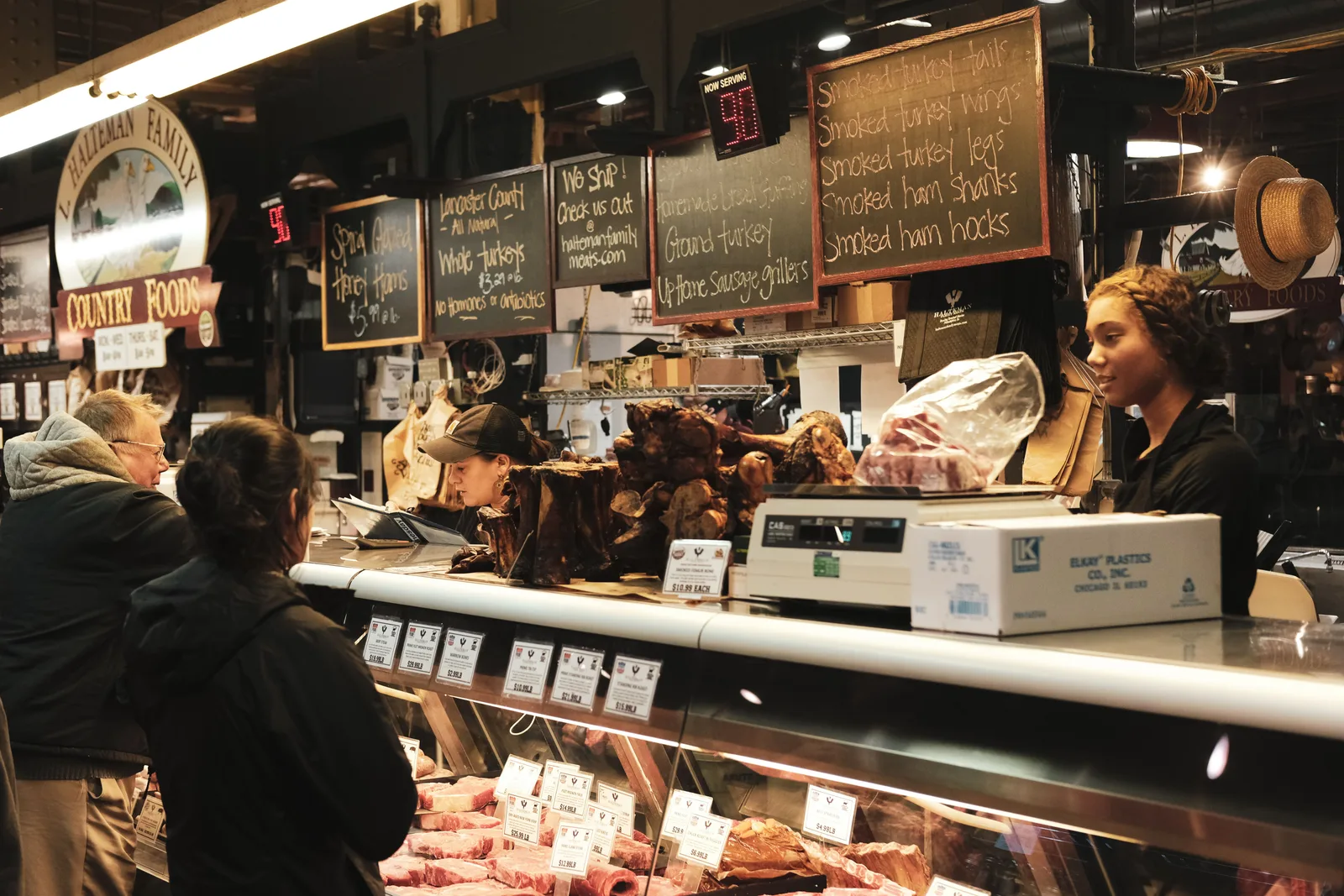 Staff serving customers across a butcher counter stocked with cuts of meat, under handwritten menu boards.
