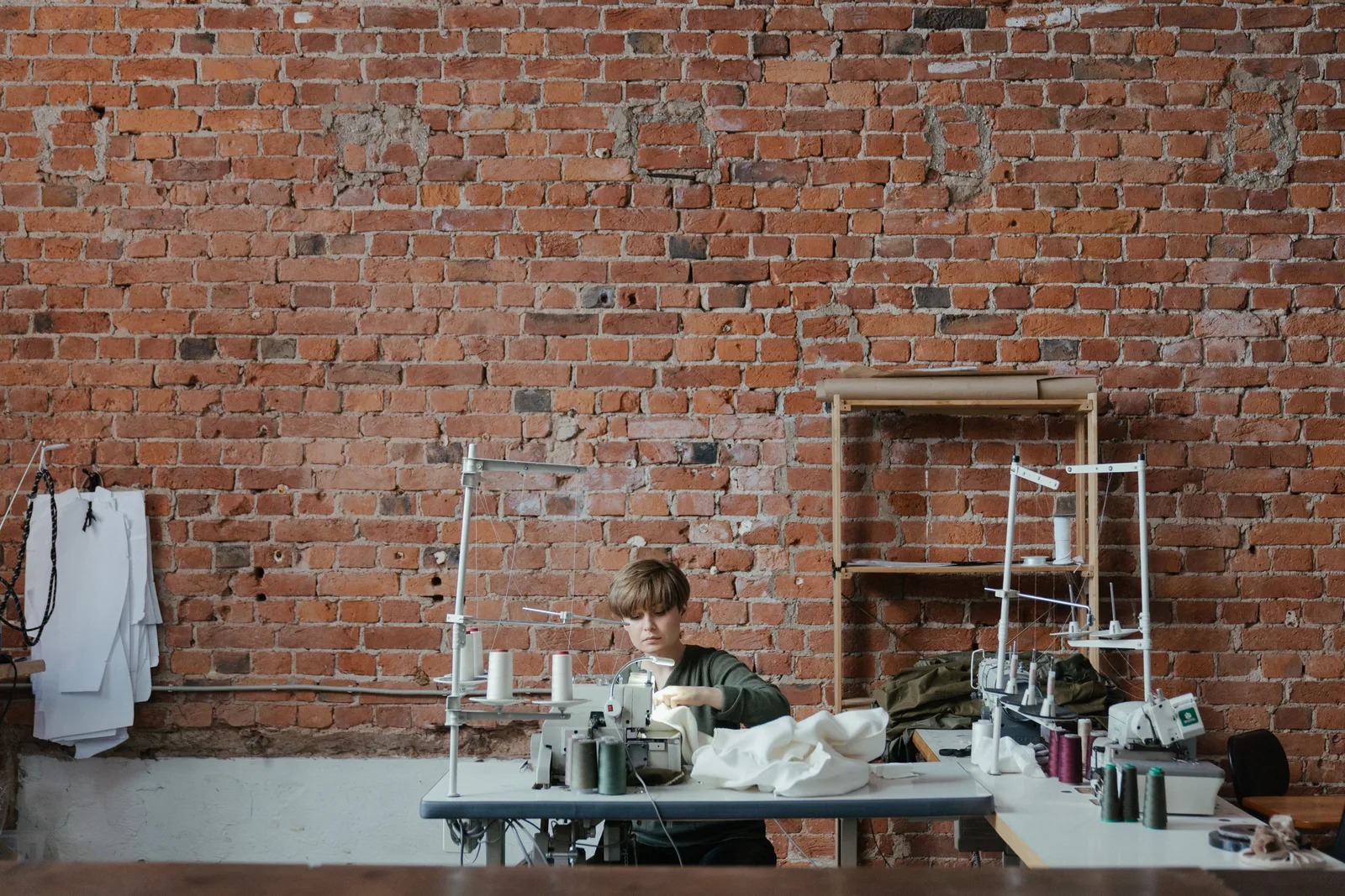 A machinist working at an industrial sewing machine in a brick walled garment workshop, with thread stands and a second machine nearby