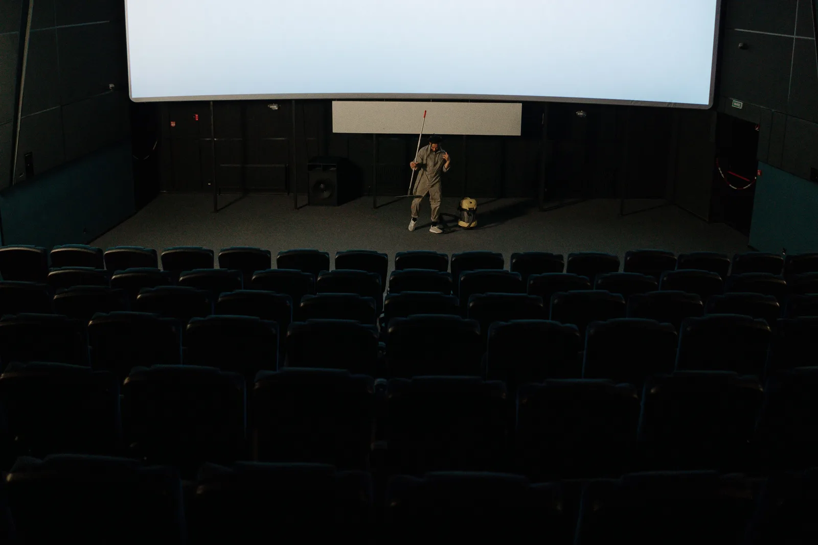 A custodian cleaning between the seat rows of an empty cinema auditorium.