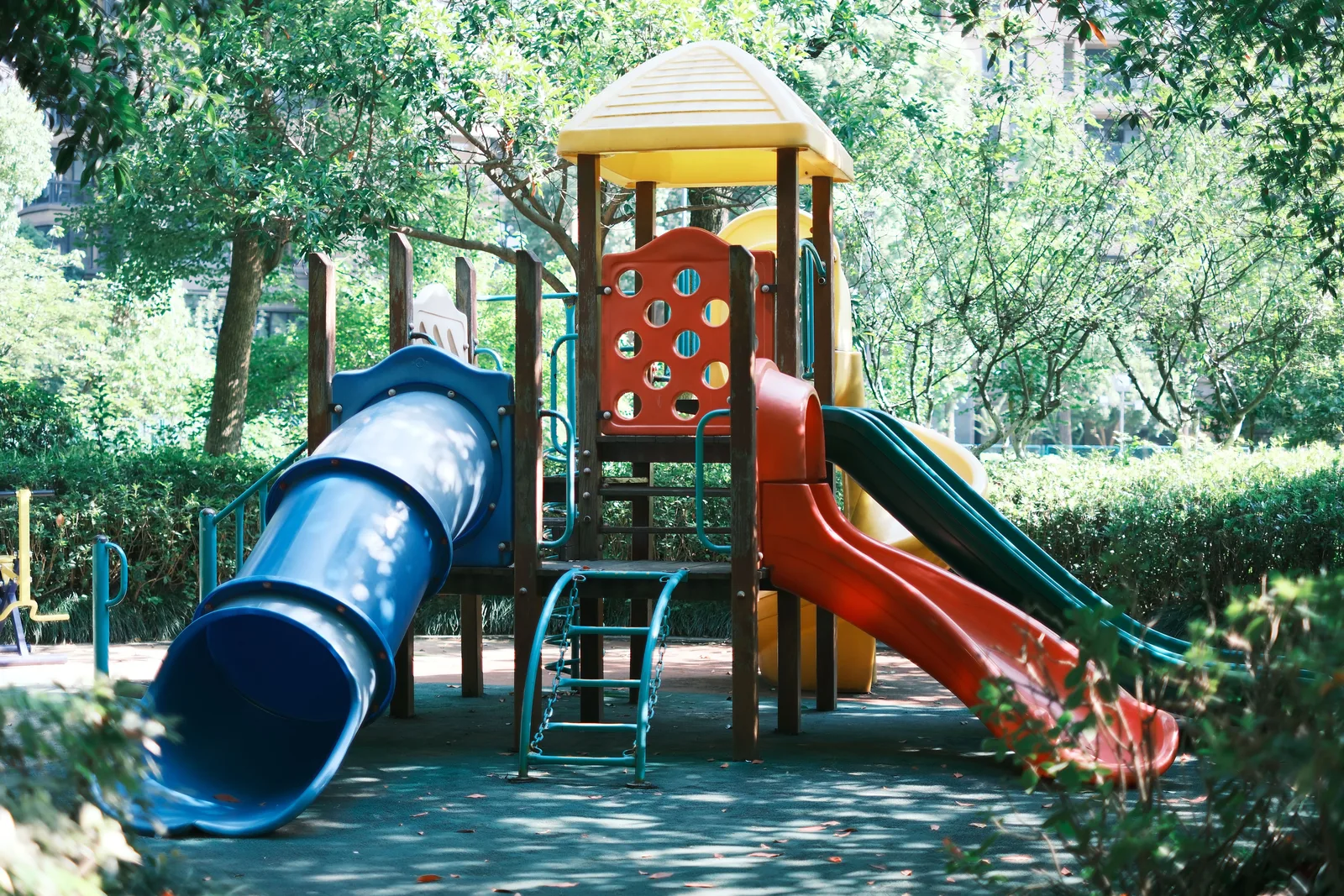 A wooden and plastic play structure with a tube slide, two open slides and a climbing ladder on a shaded rubber surfaced play area.