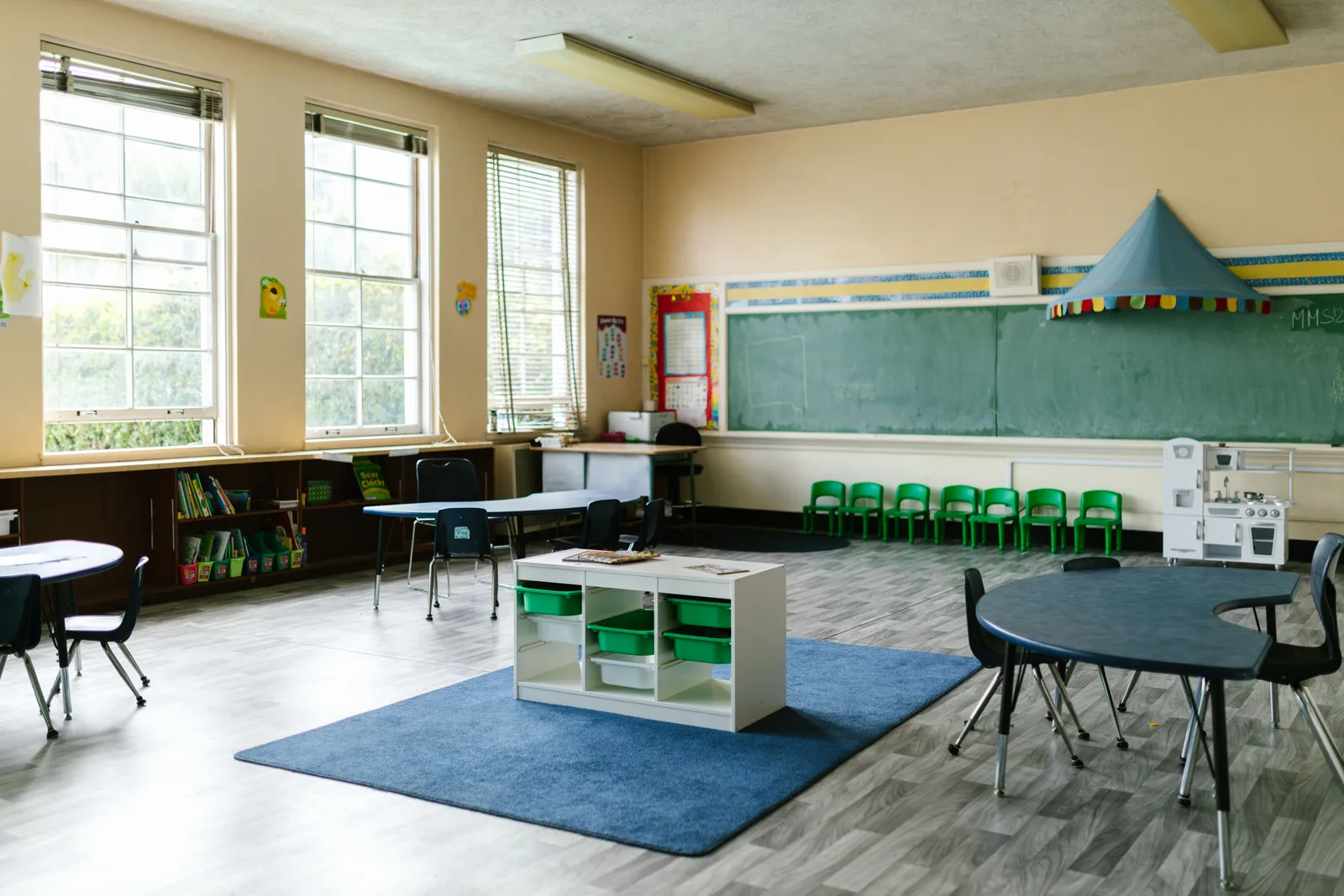 An empty early years classroom with child sized tables and chairs, a low storage unit of trays, a book shelf and a play kitchen.