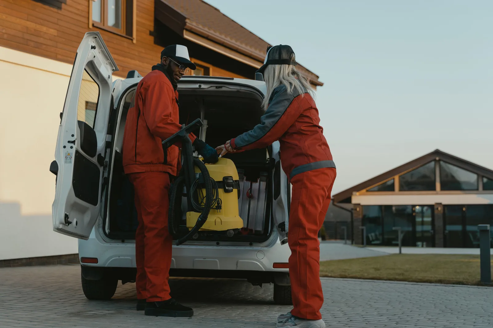 Two cleaners lifting a commercial vacuum into the back of a service van outside an office building at dusk