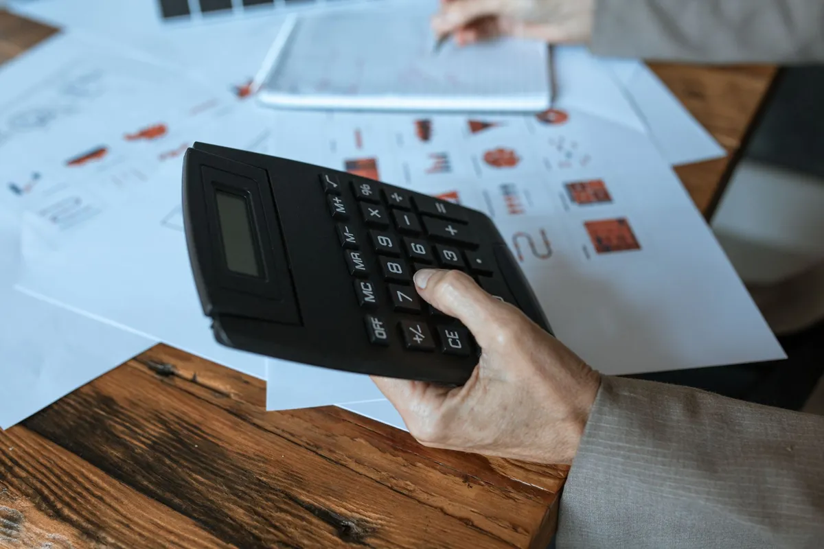 Hand holding a calculator over printed charts on a wooden desk while working out the total payback of a funding offer