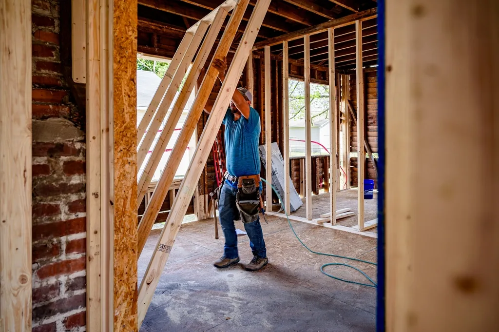 A carpenter in a work belt lifts a framed timber wall into place inside a gutted house with an exposed brick chimney