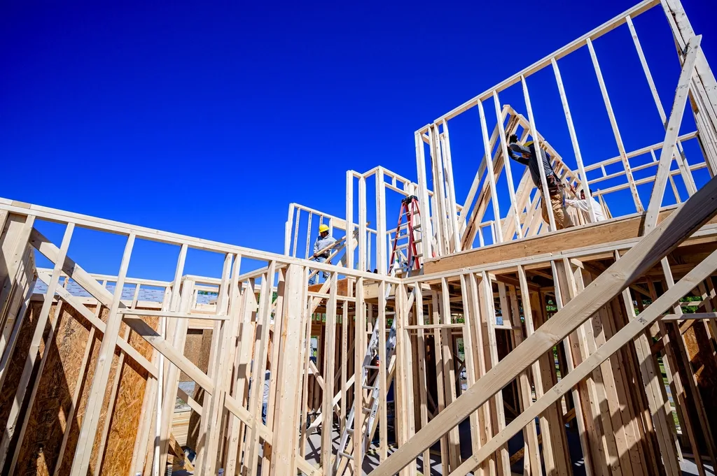 Two workers on a partly built wood-framed house, one on a stepladder fastening a wall stud, seen from below against a clear blue sky