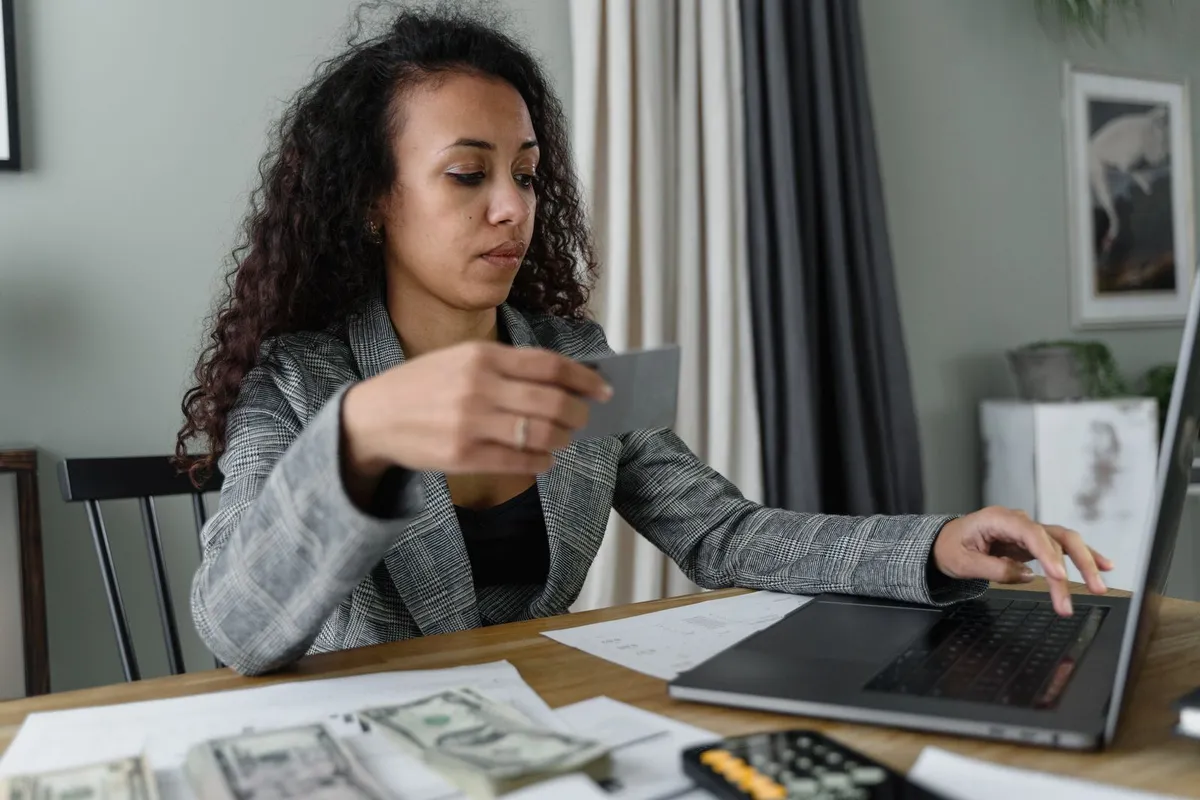 A business owner in a gray blazer holding a card with one hand and typing on a laptop with the other, with printed statements, cash and a calculator on the table in front of her