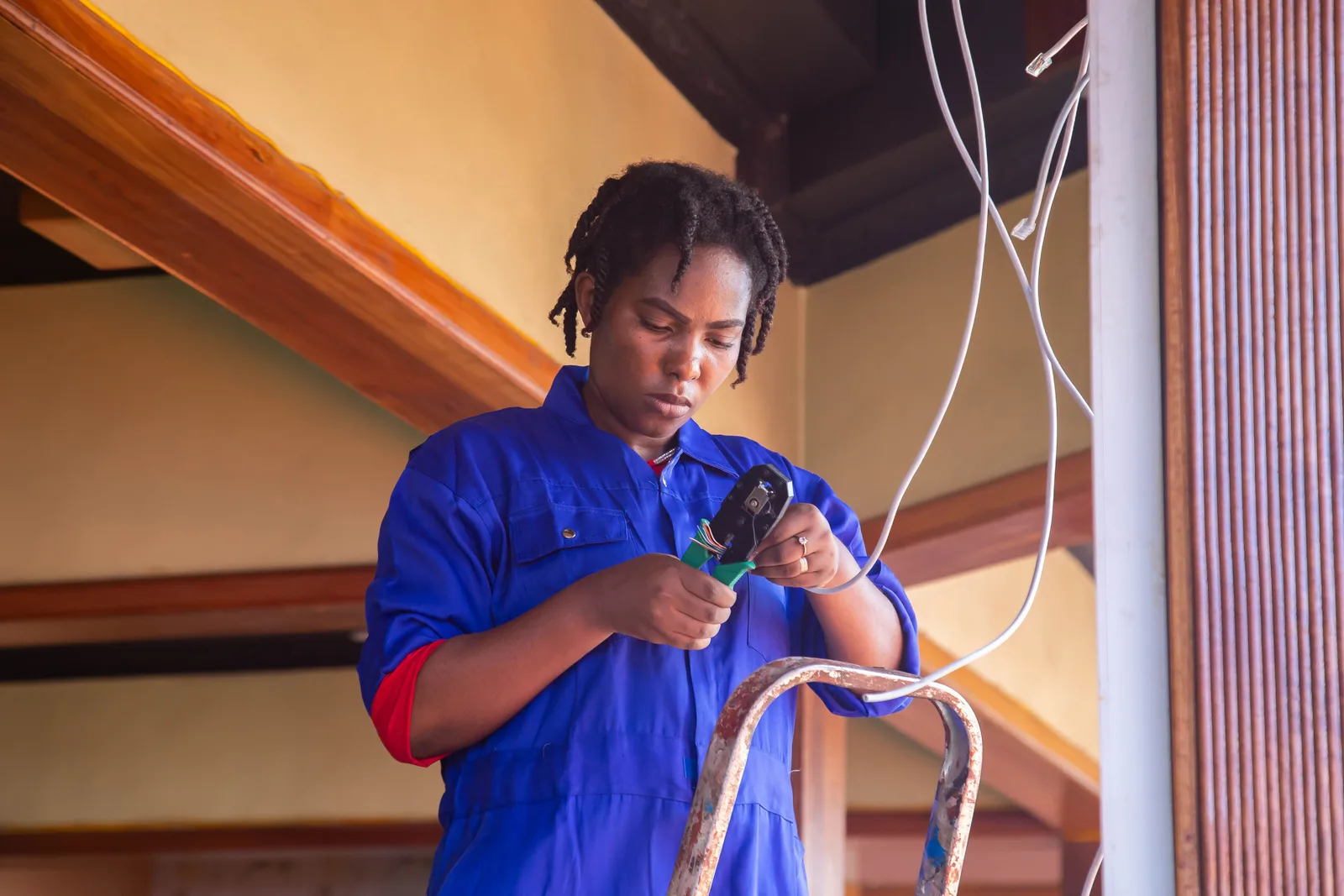 An electrician in blue coveralls stripping and terminating cable while standing on a step ladder