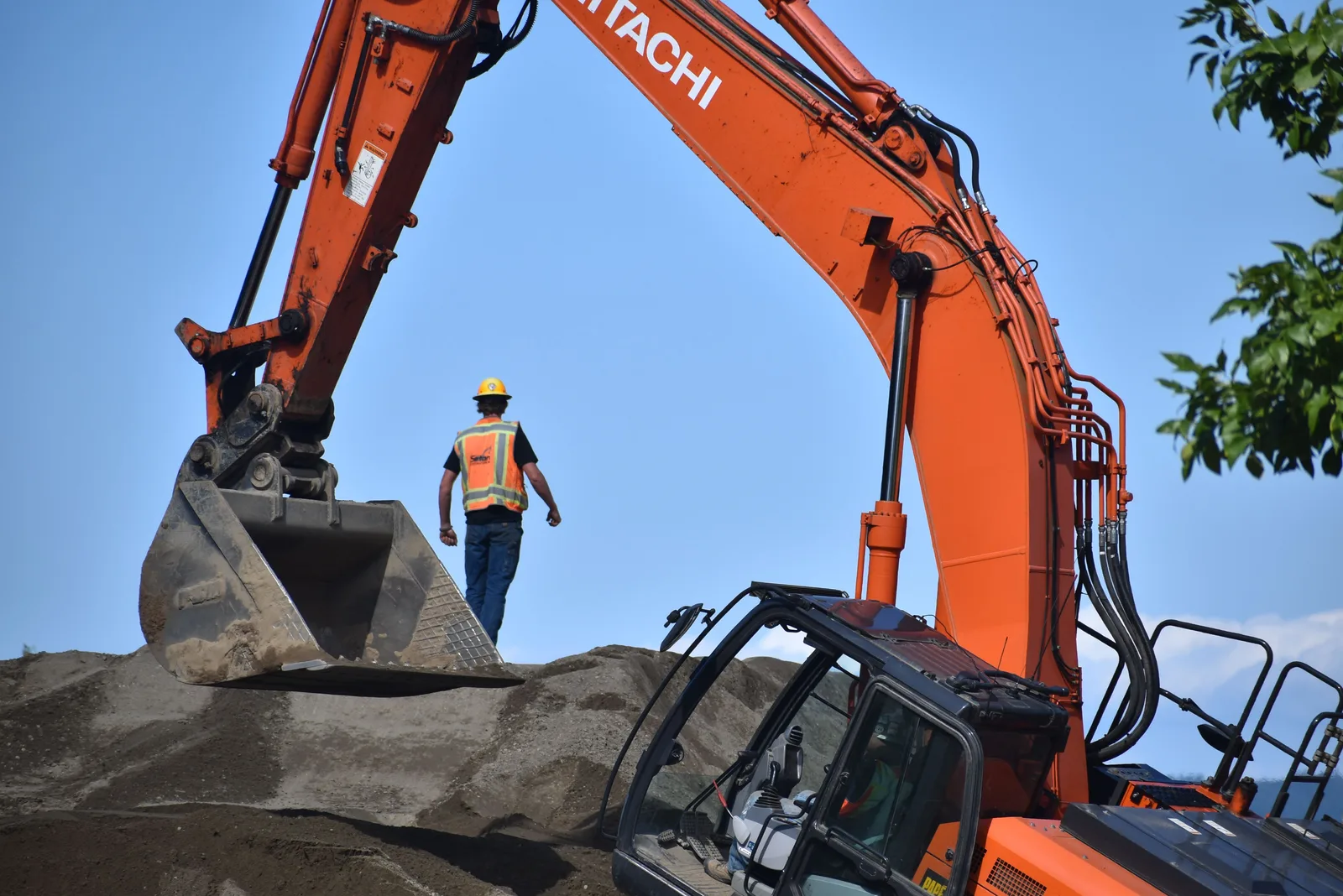 A tracked excavator with its bucket resting on a mound of earth and a worker in a hard hat standing beside it.