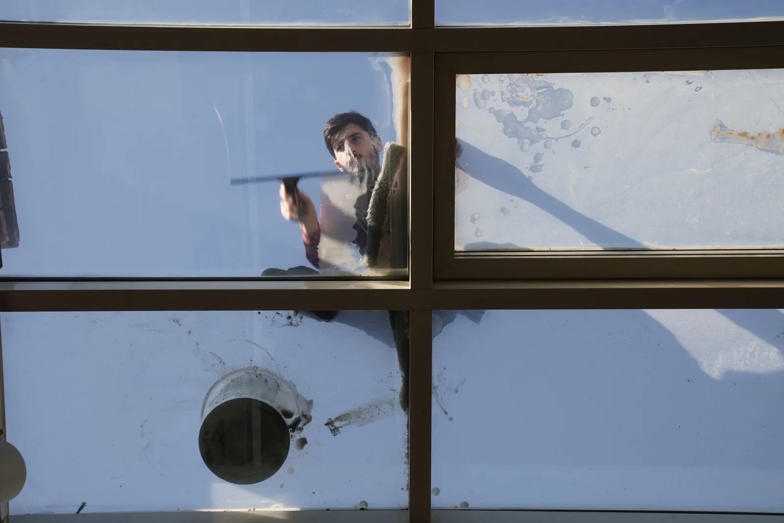 A window cleaner seen from below, drawing a squeegee across the panes of a glazed roof.