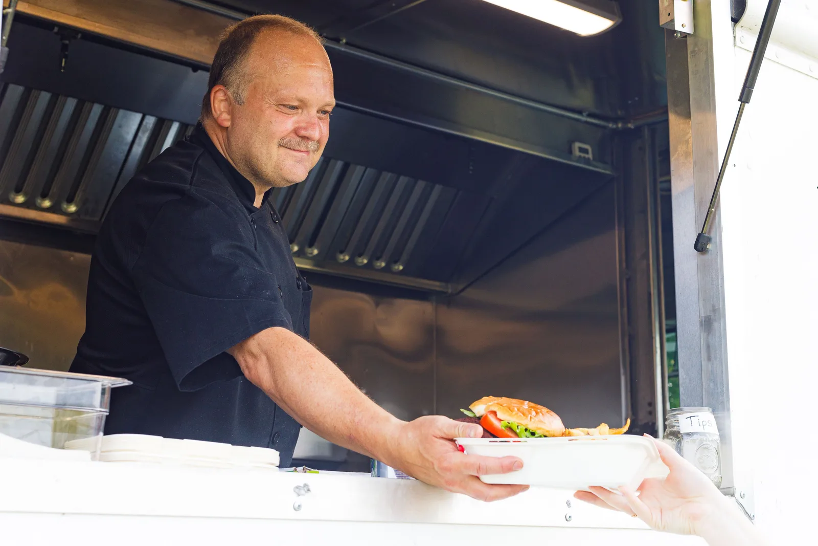 A cook in a black shirt handing a burger in a takeout box through the service window of a food truck