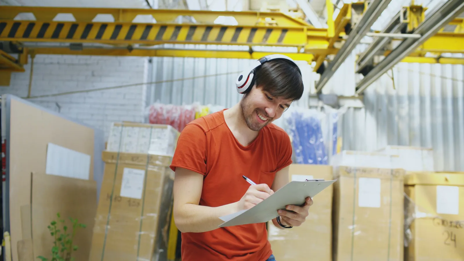 Worker in a stockroom writing on a clipboard beside stacked boxes and shelving