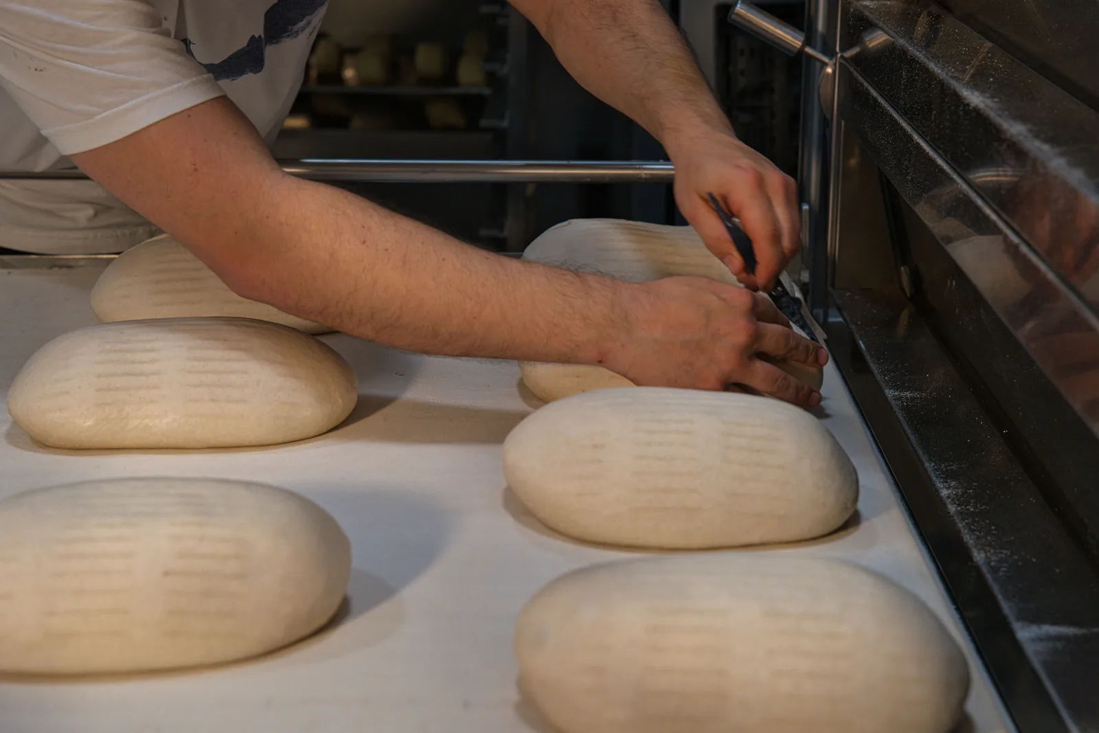 A baker's hands scoring rows of shaped dough loaves with a blade on a floured bench beside an oven