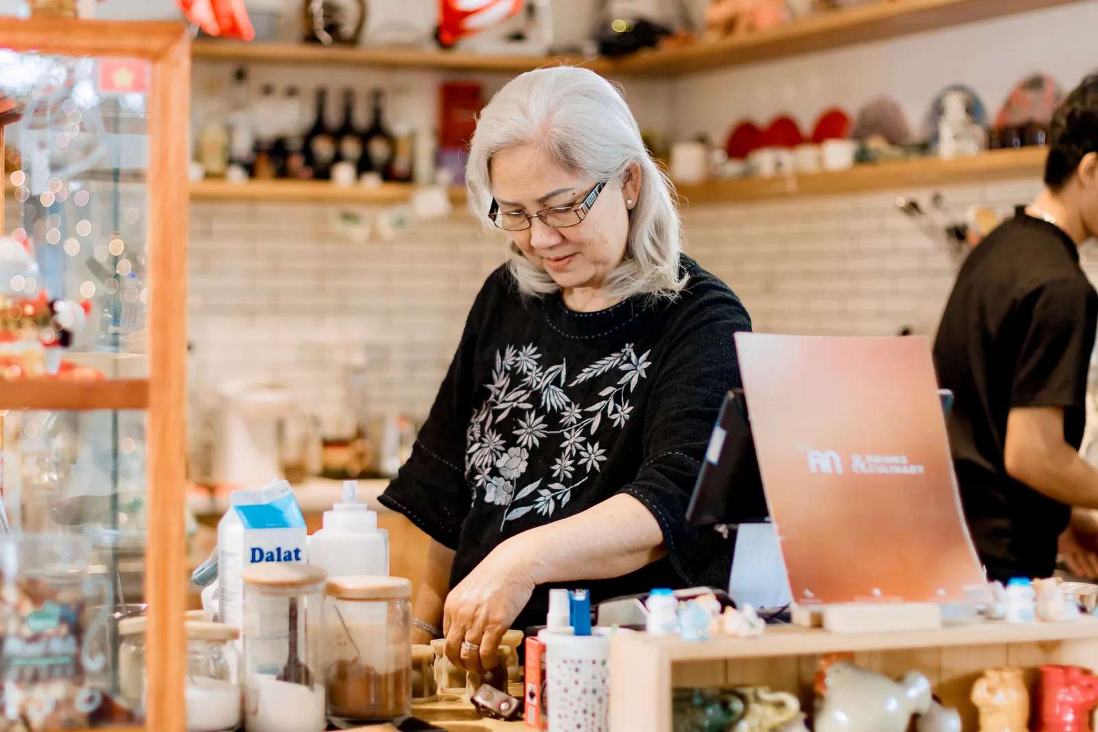 A woman in glasses working behind a small shop counter surrounded by jars, packaged goods and a card terminal, with a colleague working behind her